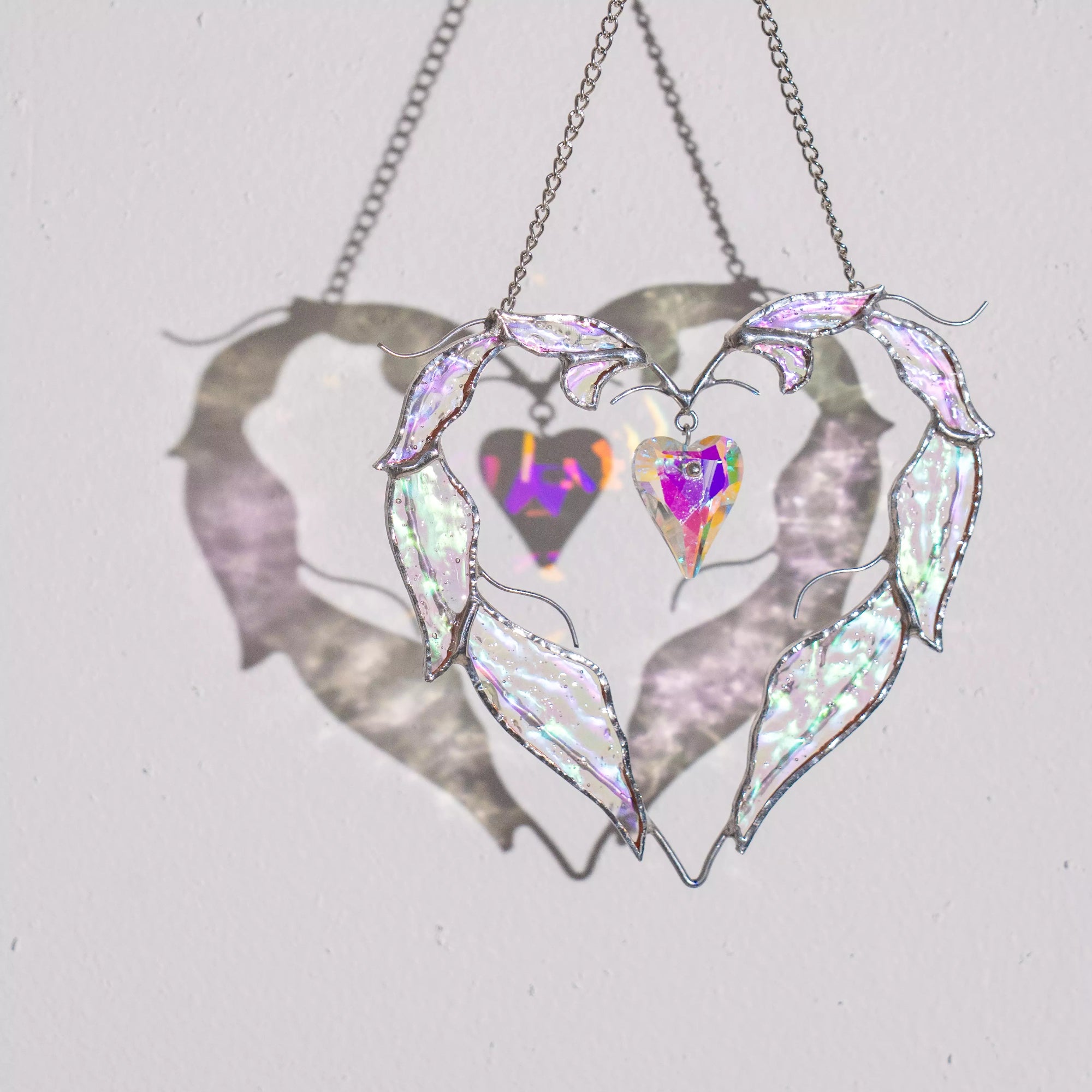 Love stained glass heart suncatcher hanging in sunlight, casting soft shadows and rainbow reflections on the wall.