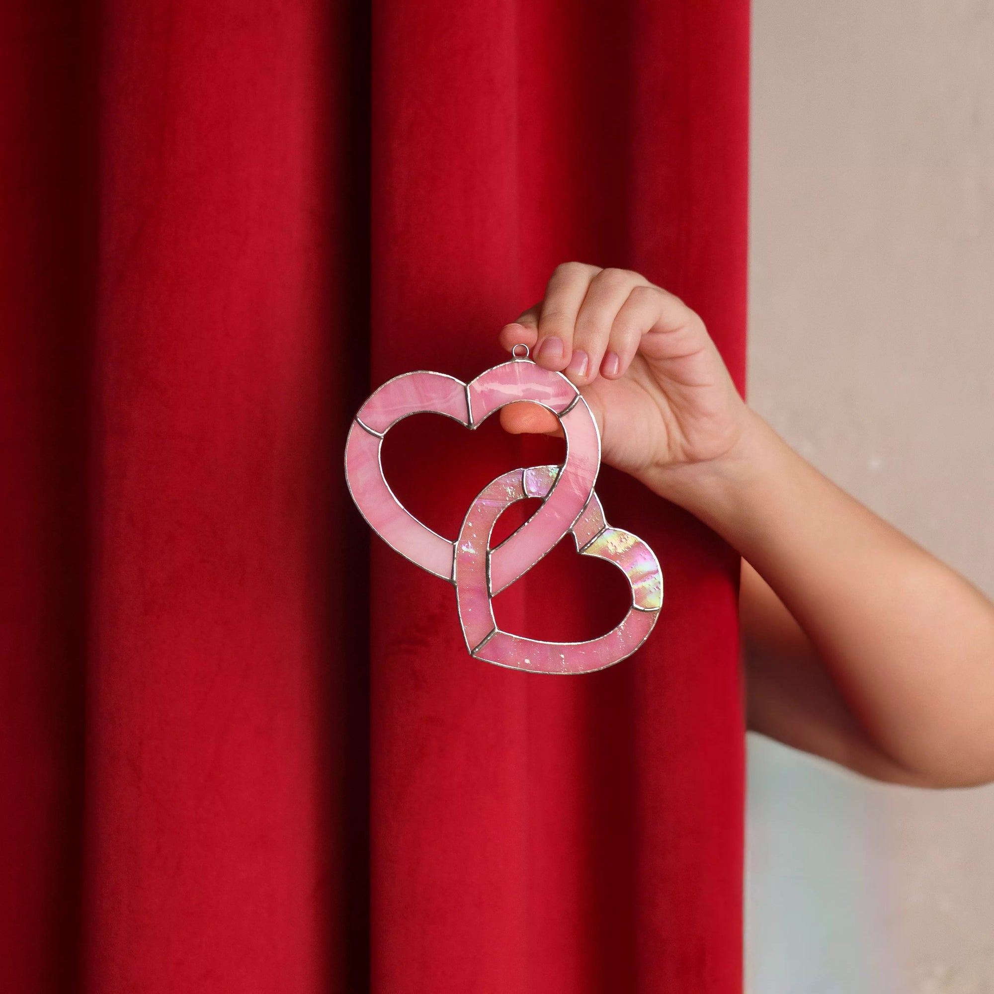 Pink stained glass heart suncatcher held against a red curtain background, highlighting romantic color contrast