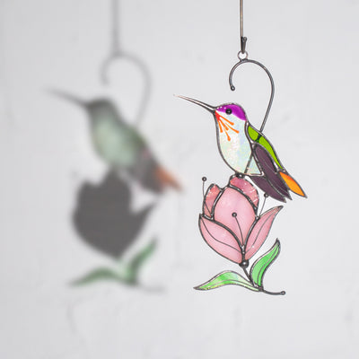 Light and shadow play behind a delicate hummingbird and pink magnolia suncatcher on a gray wall.