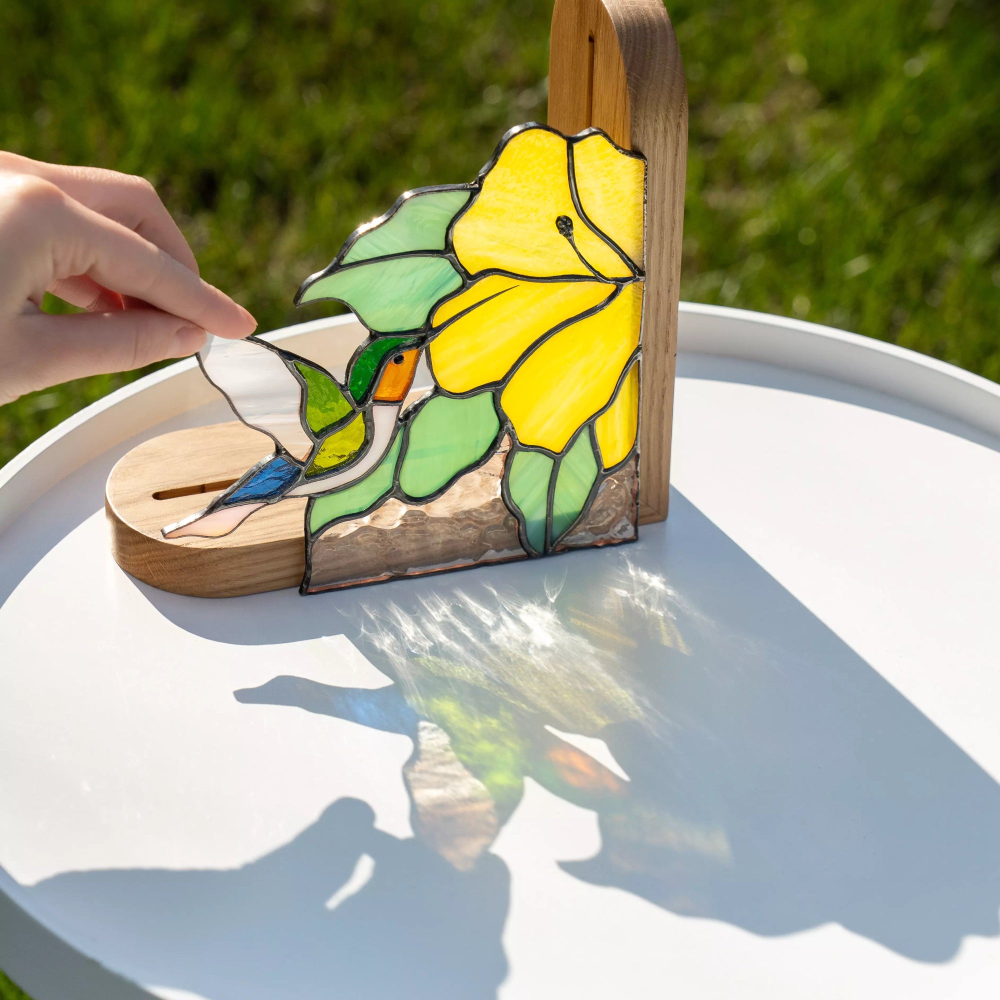 Hand holding stained glass hummingbird bookend outdoors, sunlight casting colorful shadows
