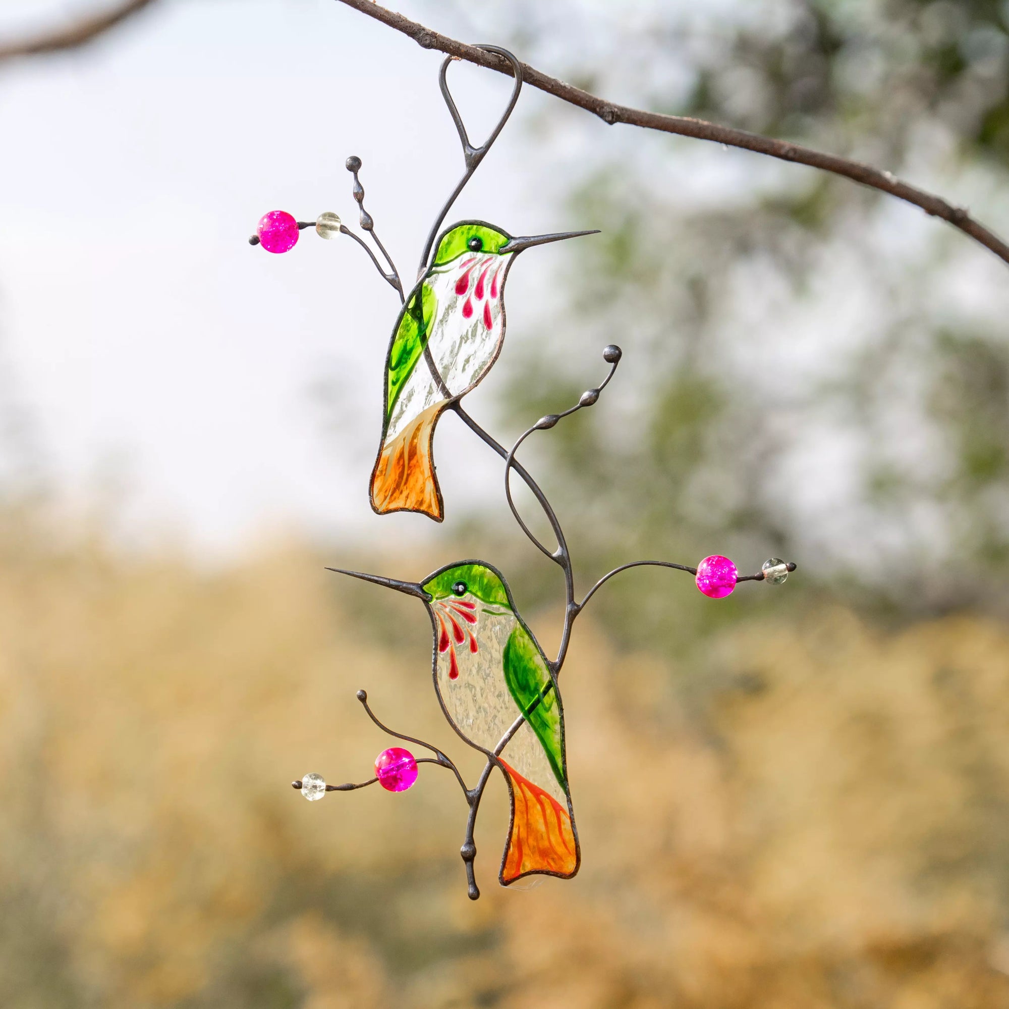 Stained glass hummingbird suncatcher on a branch with pink beads against a blurred natural background.