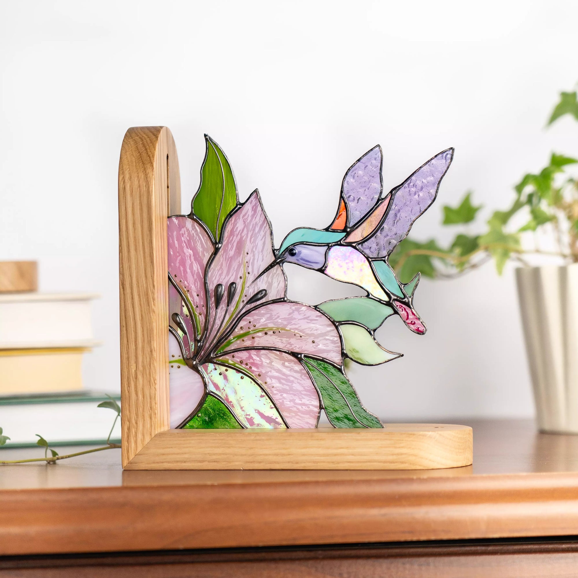 Stained glass hummingbird bookend with pink flower on wooden table near books and plant