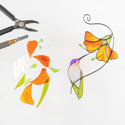 Glass cutting tools and pieces beside completed stained glass hummingbird and orange flower