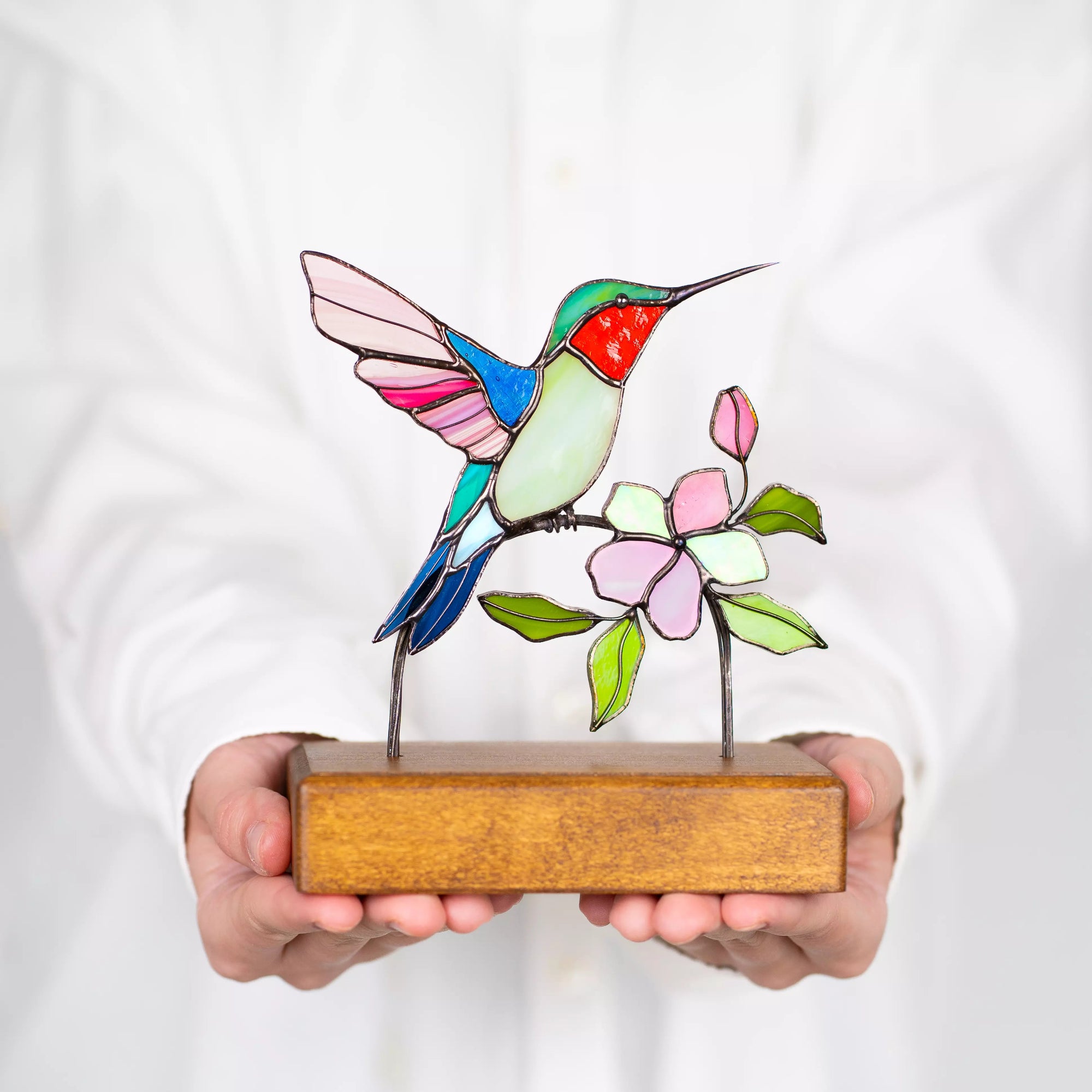 Hands holding stained glass hummingbird sculpture against a white background