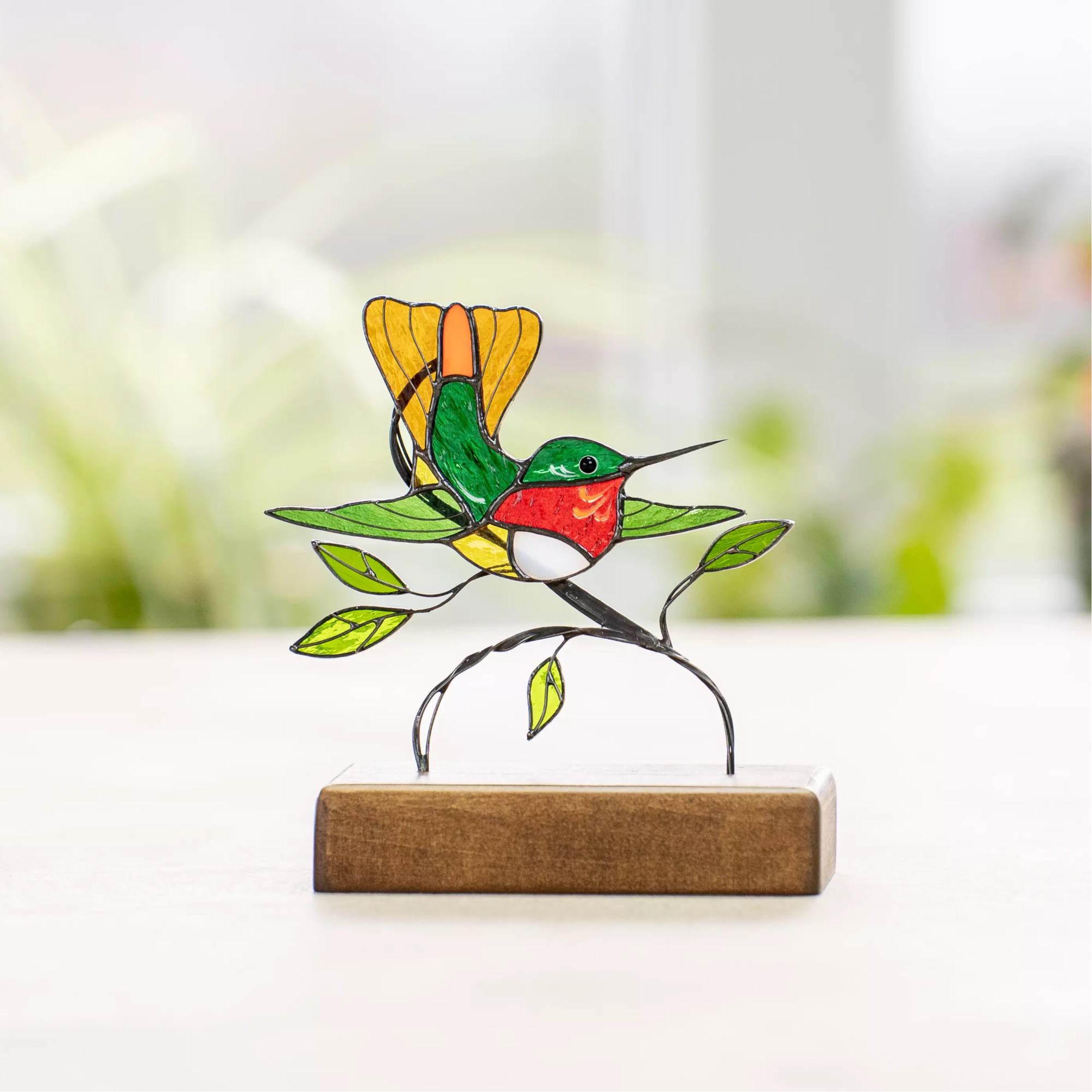 Hummingbird suncatcher made of stained glass, resting on a wooden base with leafy metal details and natural light in the background.