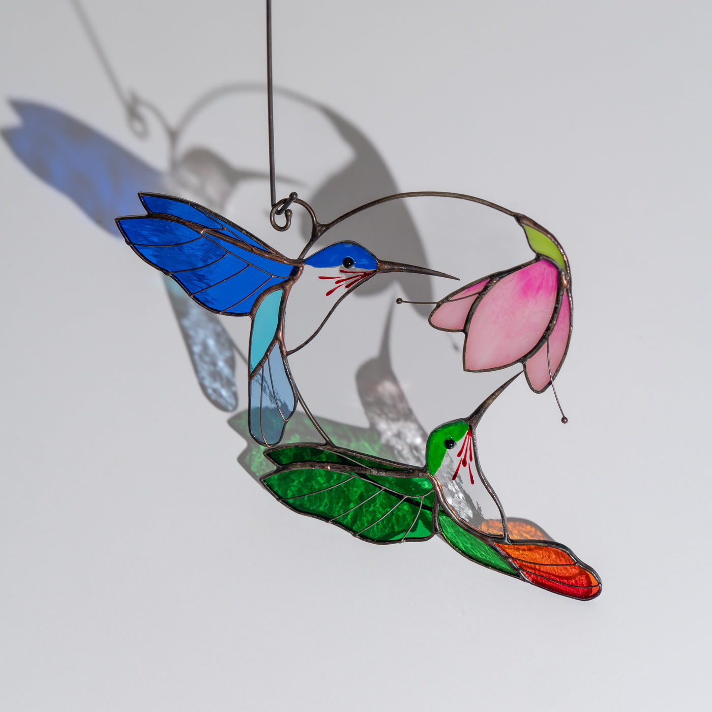 Hanging stained glass with green and blue hummingbirds flying toward a pink flower