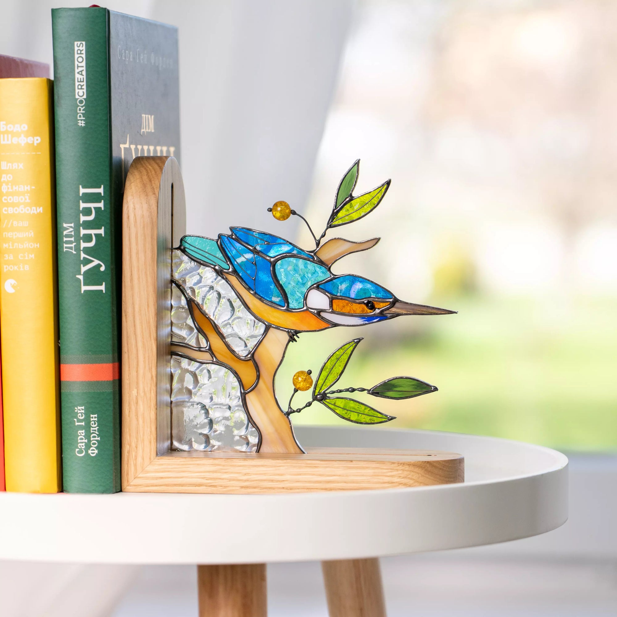 Handcrafted stained glass kingfisher bird bookend in blue and orange colors. Features intricate solder lines, green glass leaves, and amber-colored beads on a natural wooden base.