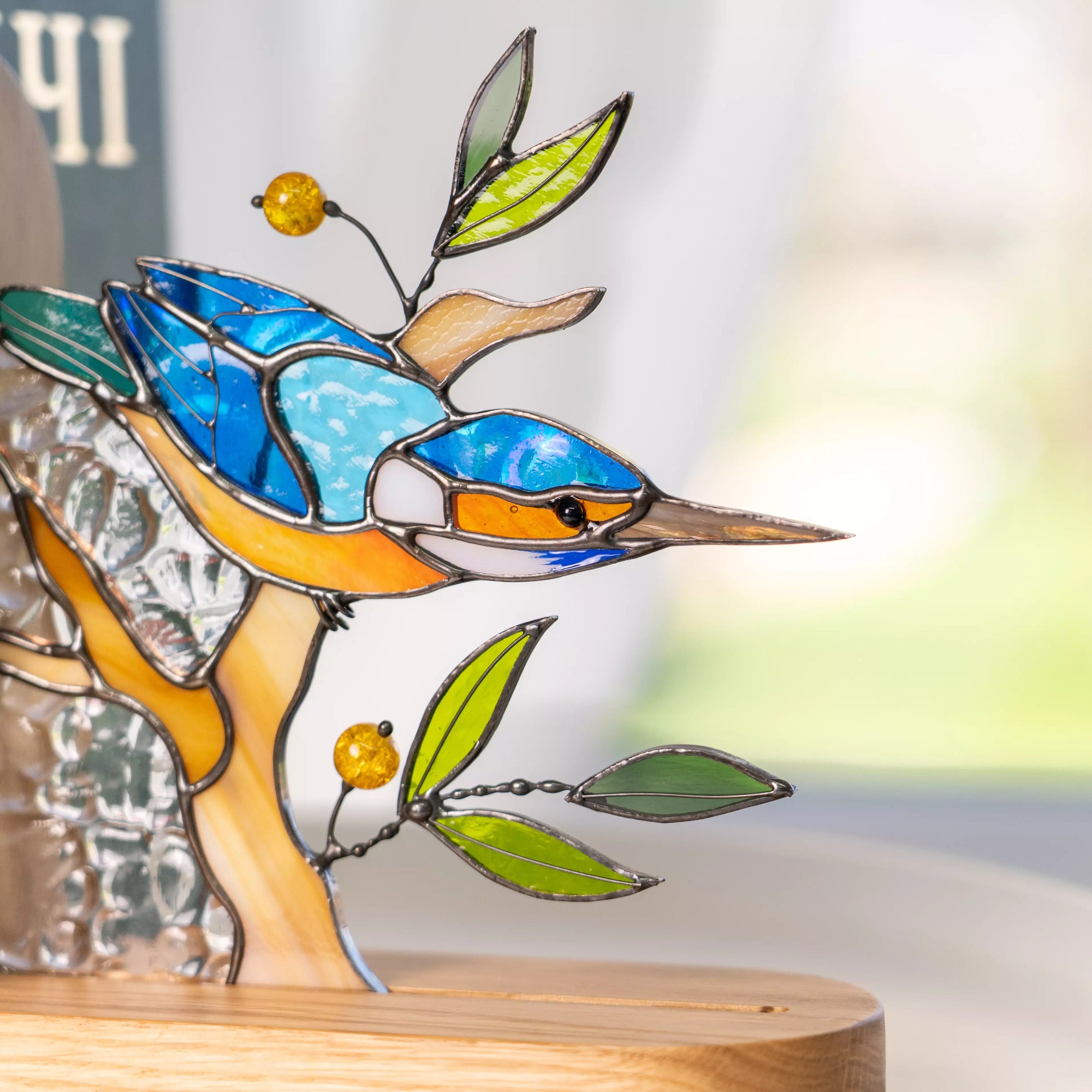 Handcrafted stained glass kingfisher bird bookend in blue and orange colors. Features intricate solder lines, green glass leaves, and amber-colored beads on a natural wooden base.