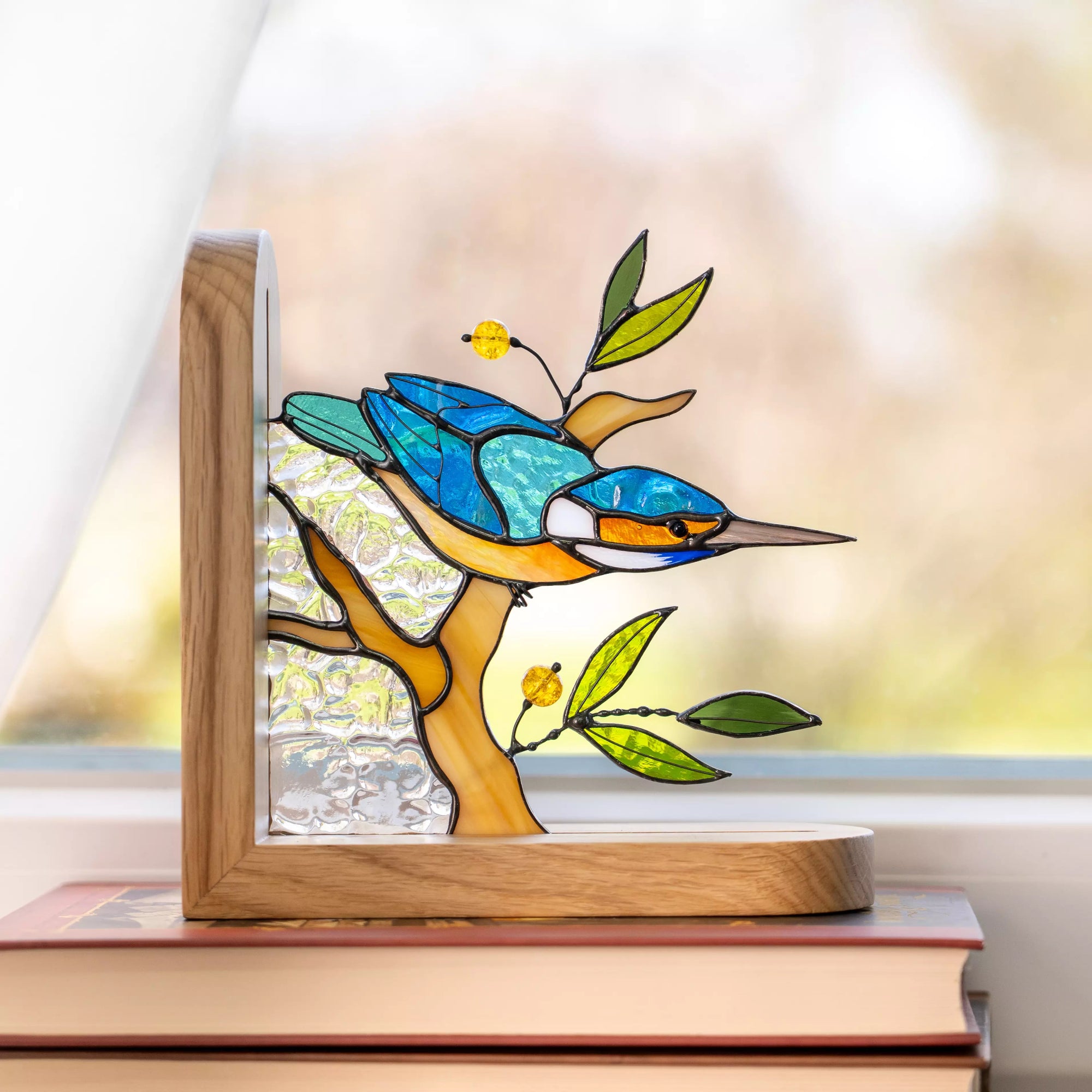 Handcrafted stained glass kingfisher bookend sitting on a stack of books by a window. Natural sunlight illuminates the textured glass, highlighting the bright blue and orange colors