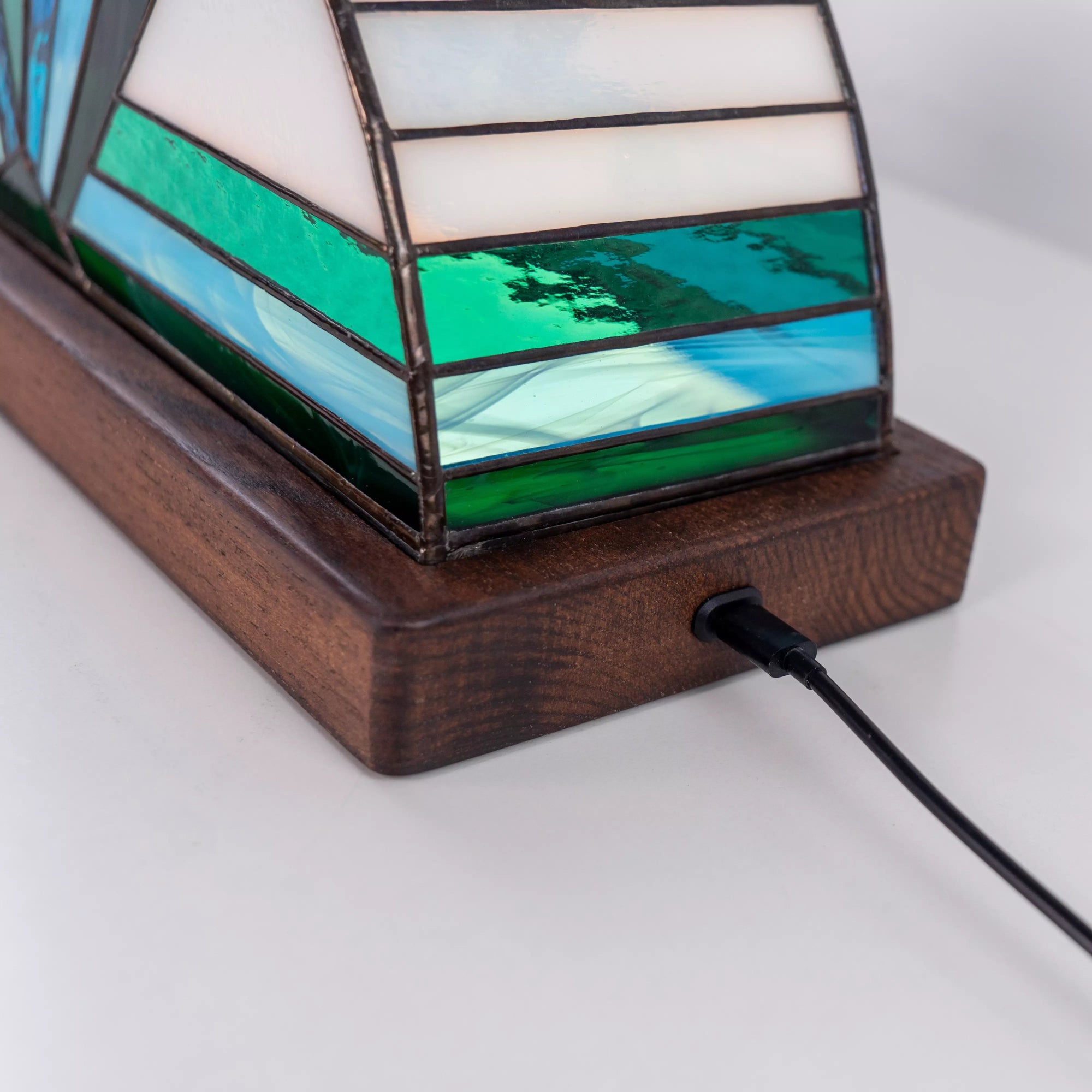 Close-up of stained glass lamp base showing blue and green glass details on a dark wooden stand with power connection, handmade bedside lighting decor.