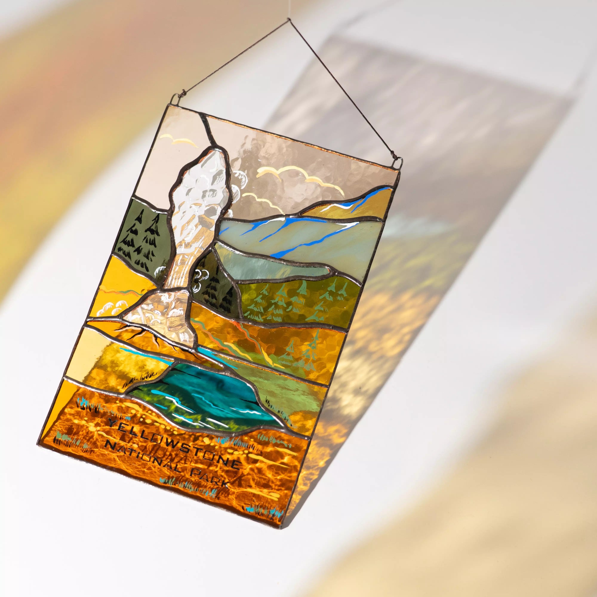 A Yellowstone geyser stained glass panel casting a warm amber and blue shadow on a white wall.