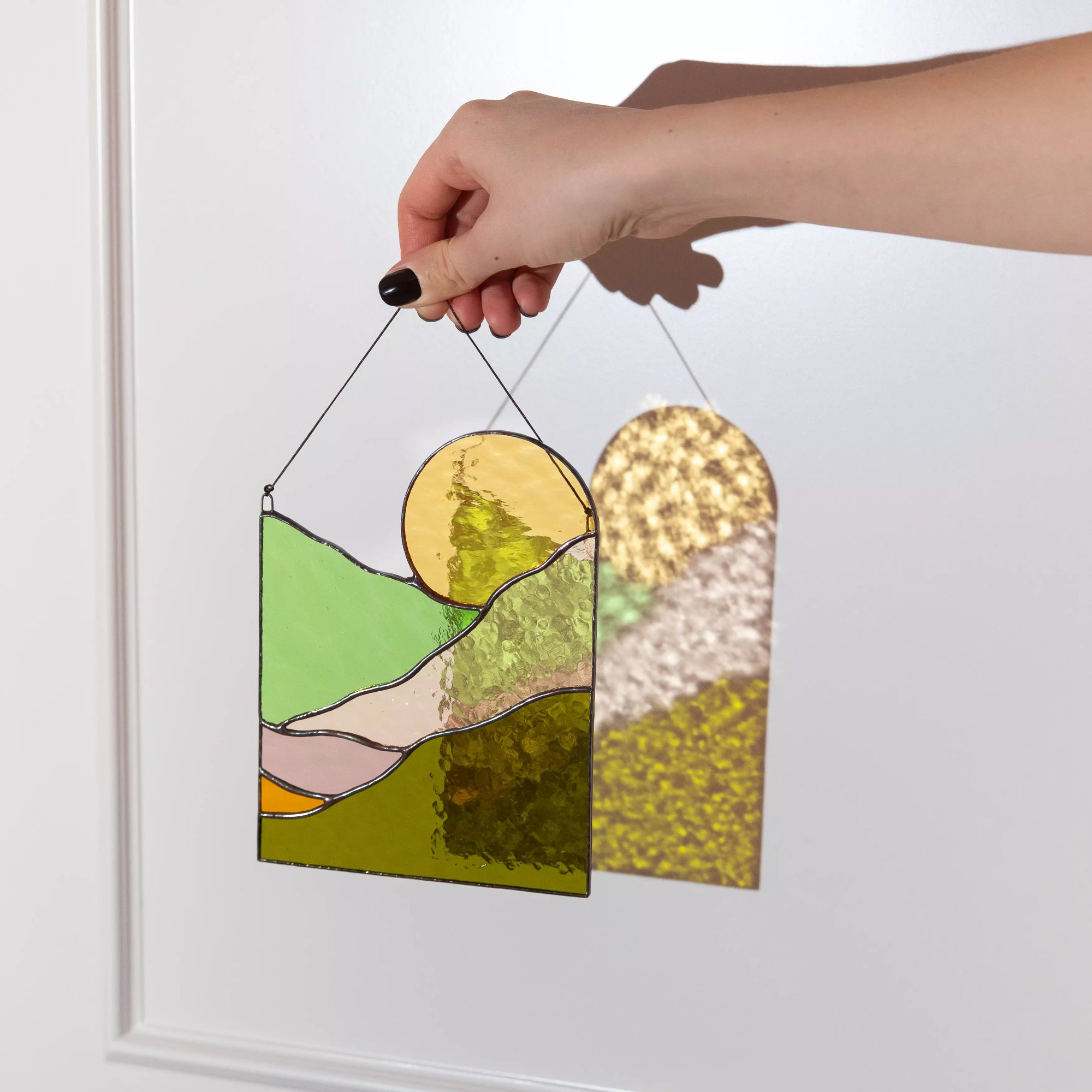 A person holding a sun landscape stained glass suncatcher that casts a warm, textured shadow.