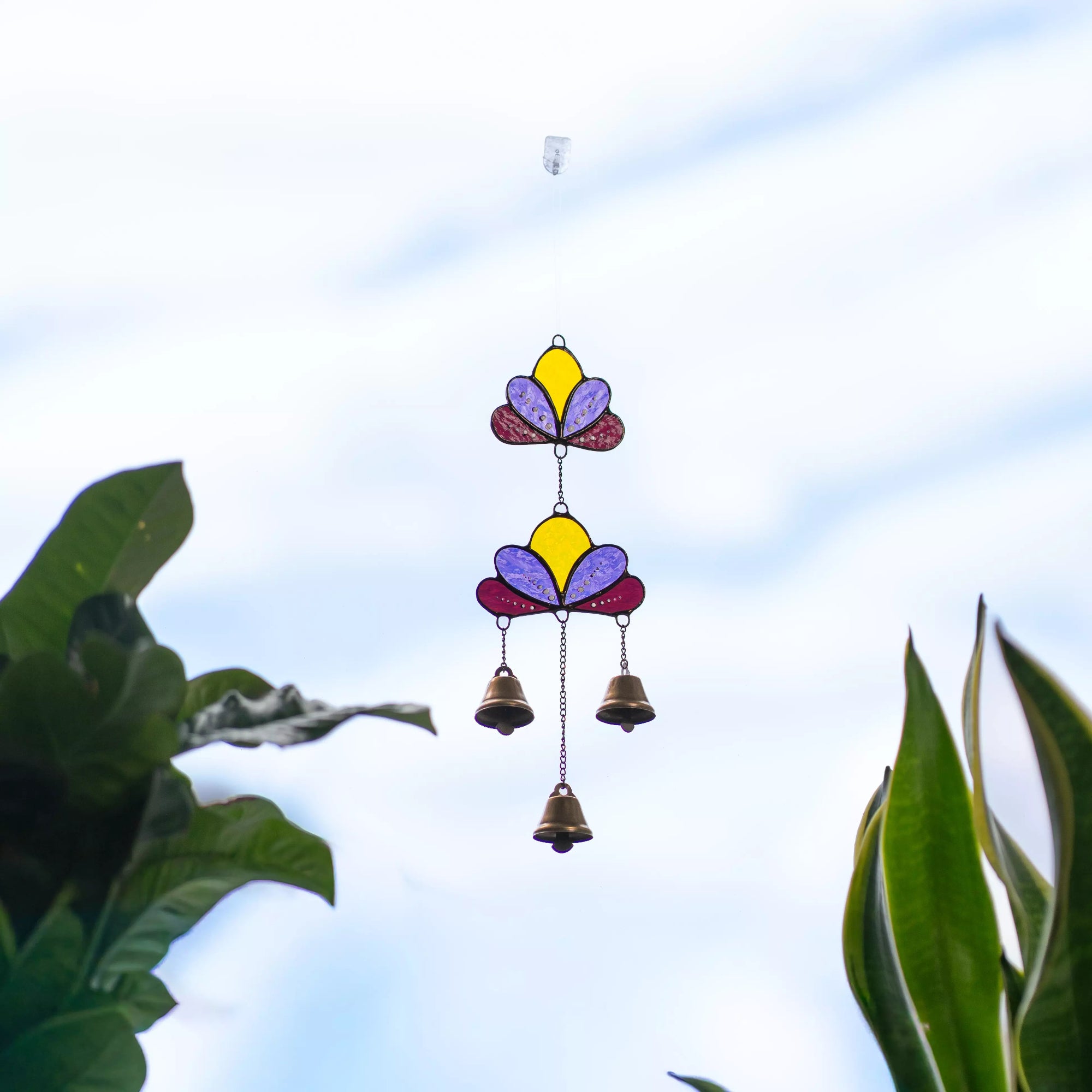 A person holds a stained glass lotus wind chime with small brass bells in front of a window filled with green house plants.