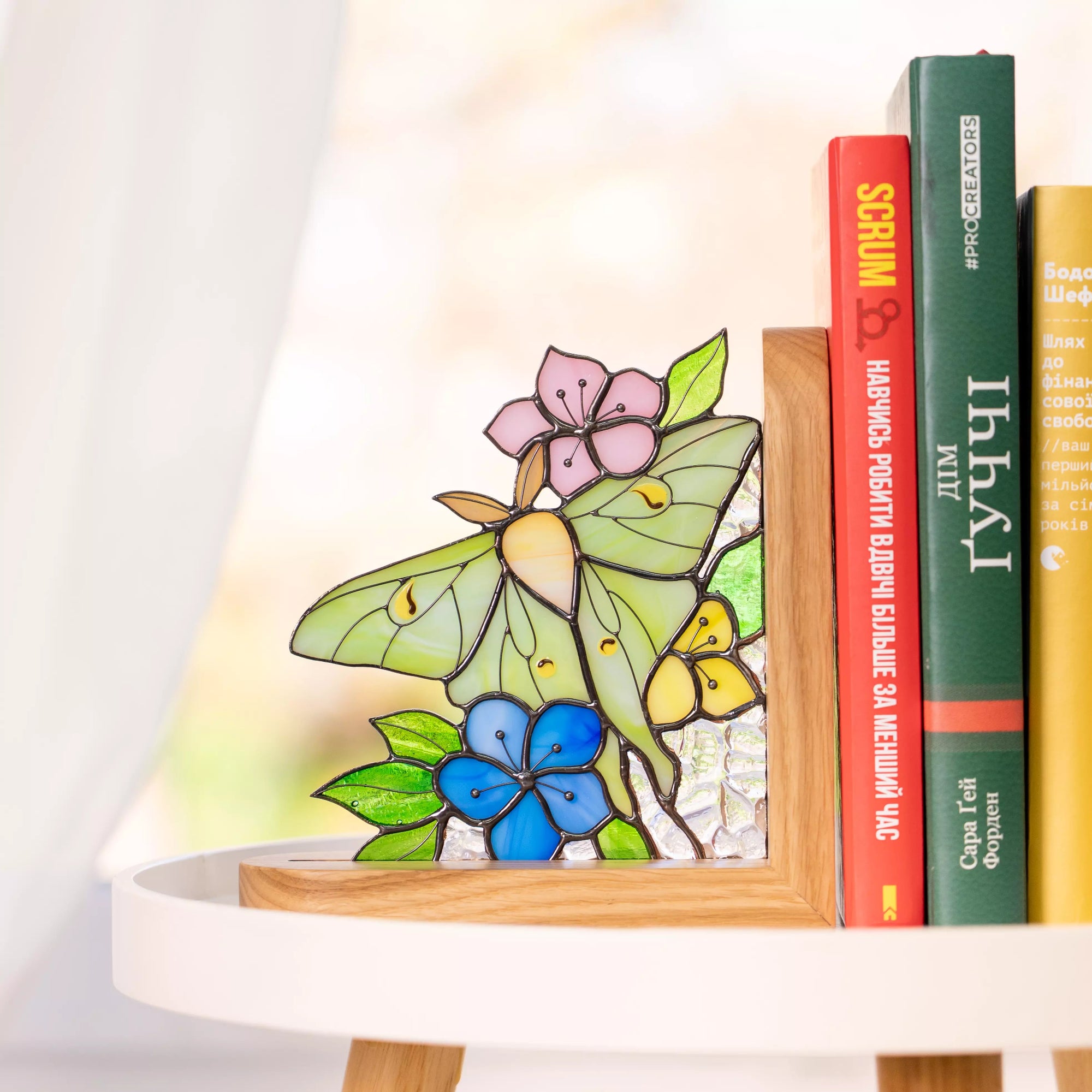 Stained glass bookend luna moth with green wings, pink and blue flowers, displayed next to books on a white shelf