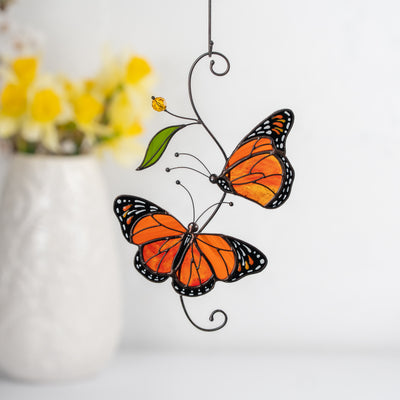 Two stained glass monarch butterflies in vibrant orange shades with a green leaf accent, hanging near a white vase with yellow flowers.