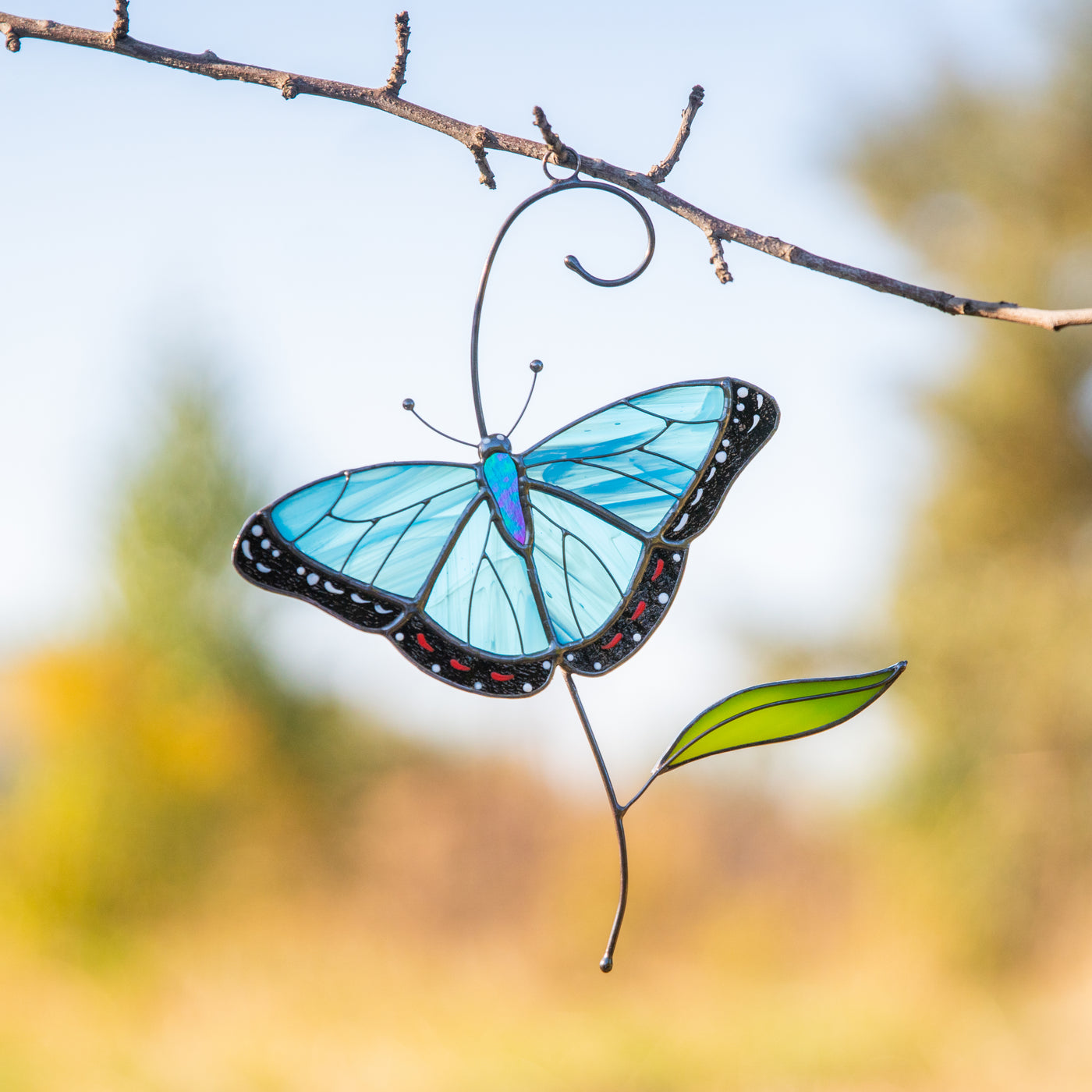 A stained glass morpho butterfly suncatcher with vibrant blue wings and green leaf hanging on the branch against a sunny garden background.