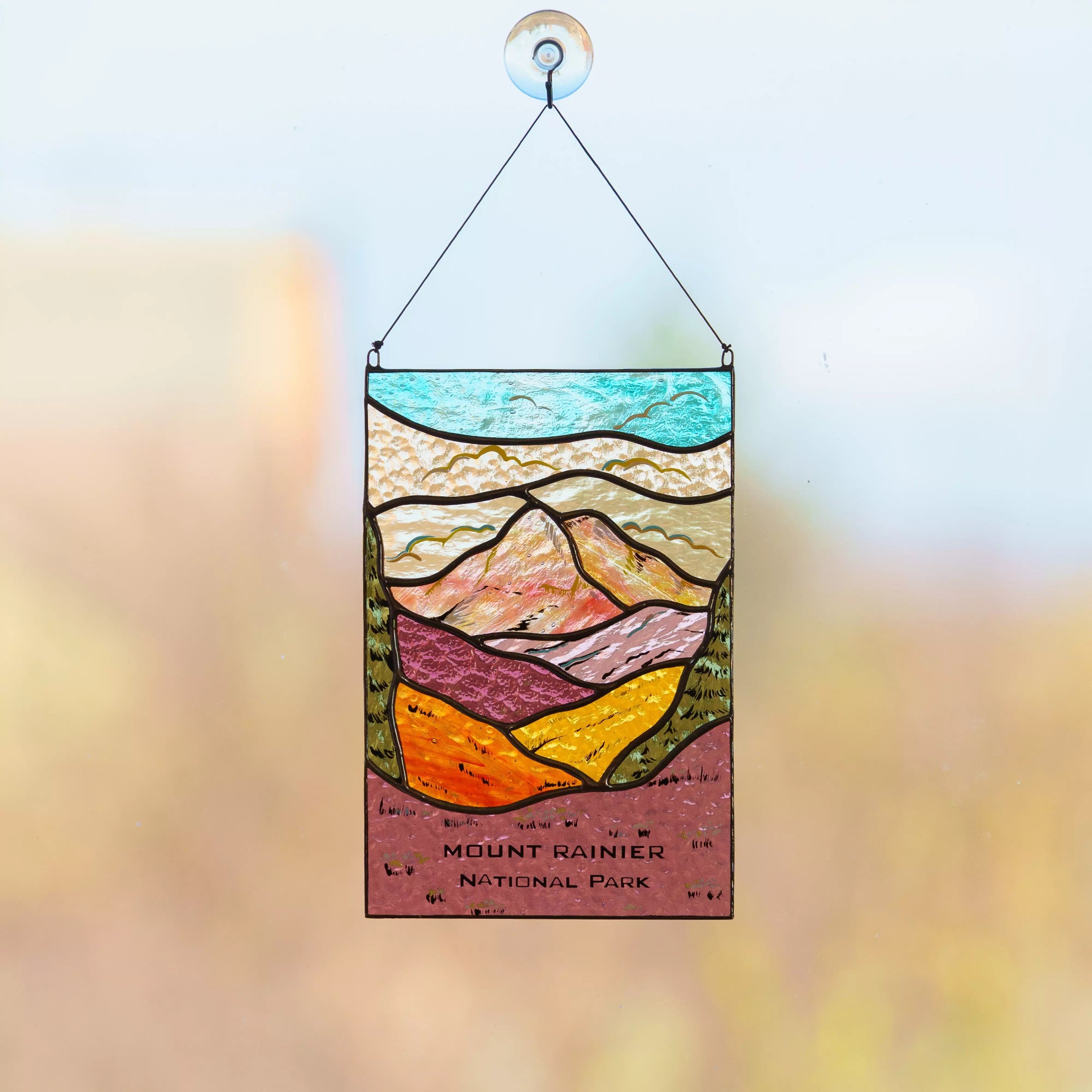 A detailed Mount Rainier National Park stained glass panel hanging against a soft, sunlit outdoor background.