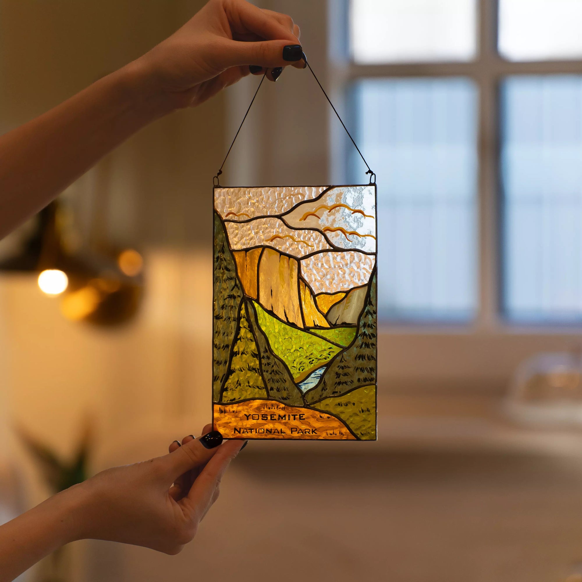 A person holding the Yosemite National Park stained glass panel in a warm cozy interior setting near a window.