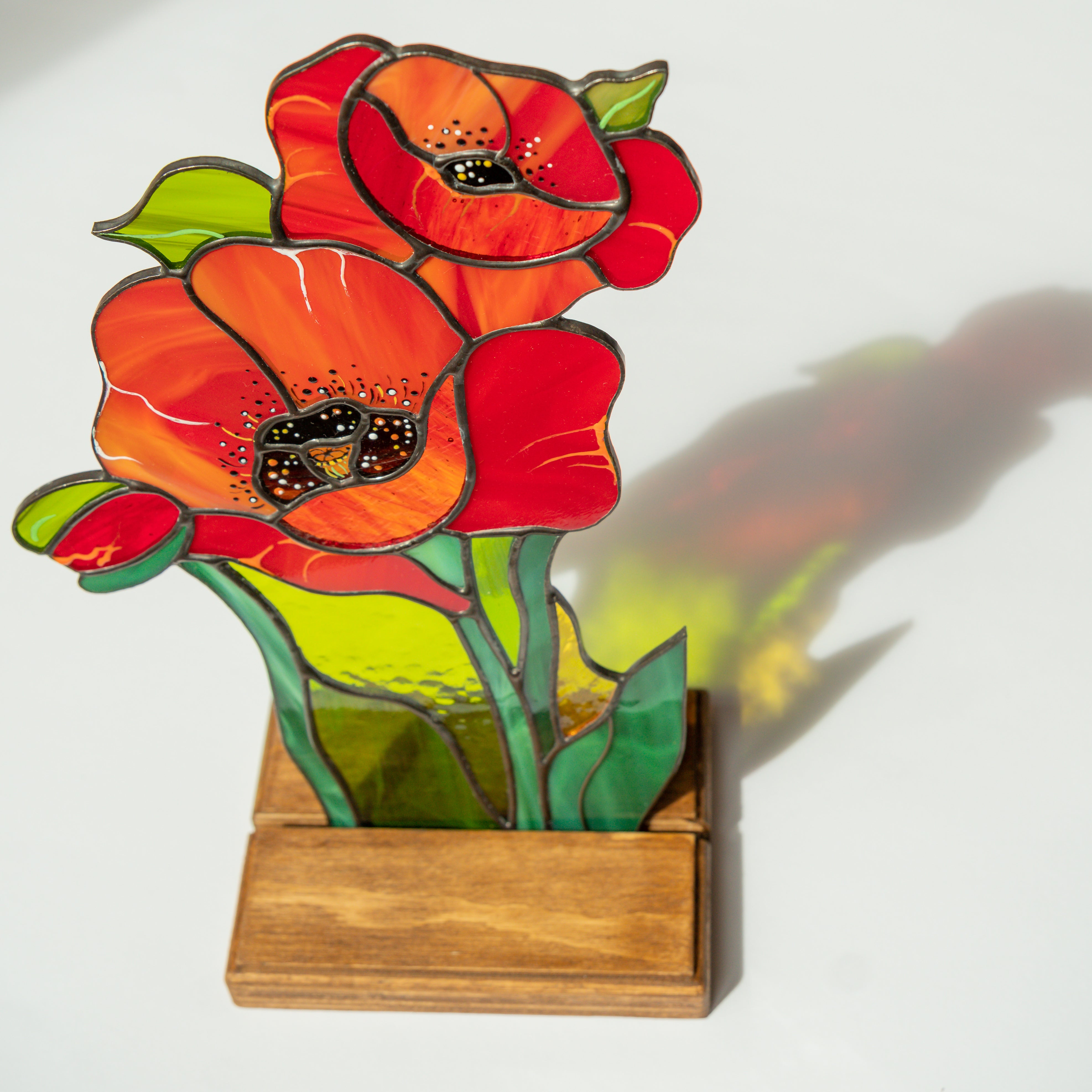 Two red-orange stained glass poppies in a wooden base casting colorful shadows on a white surface.
