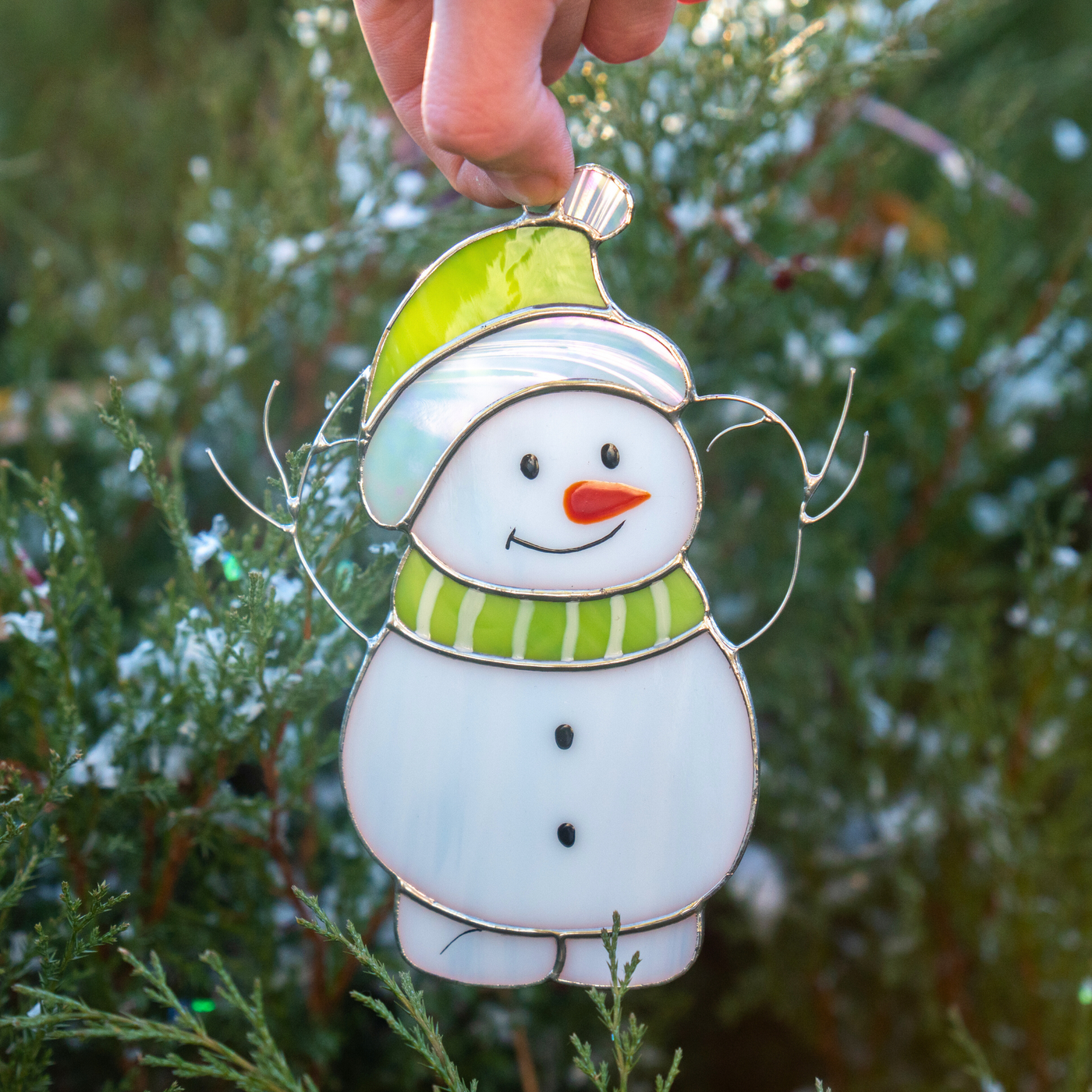 A handmade stained glass snowman with a green hat and striped scarf, held outdoors among green branches.