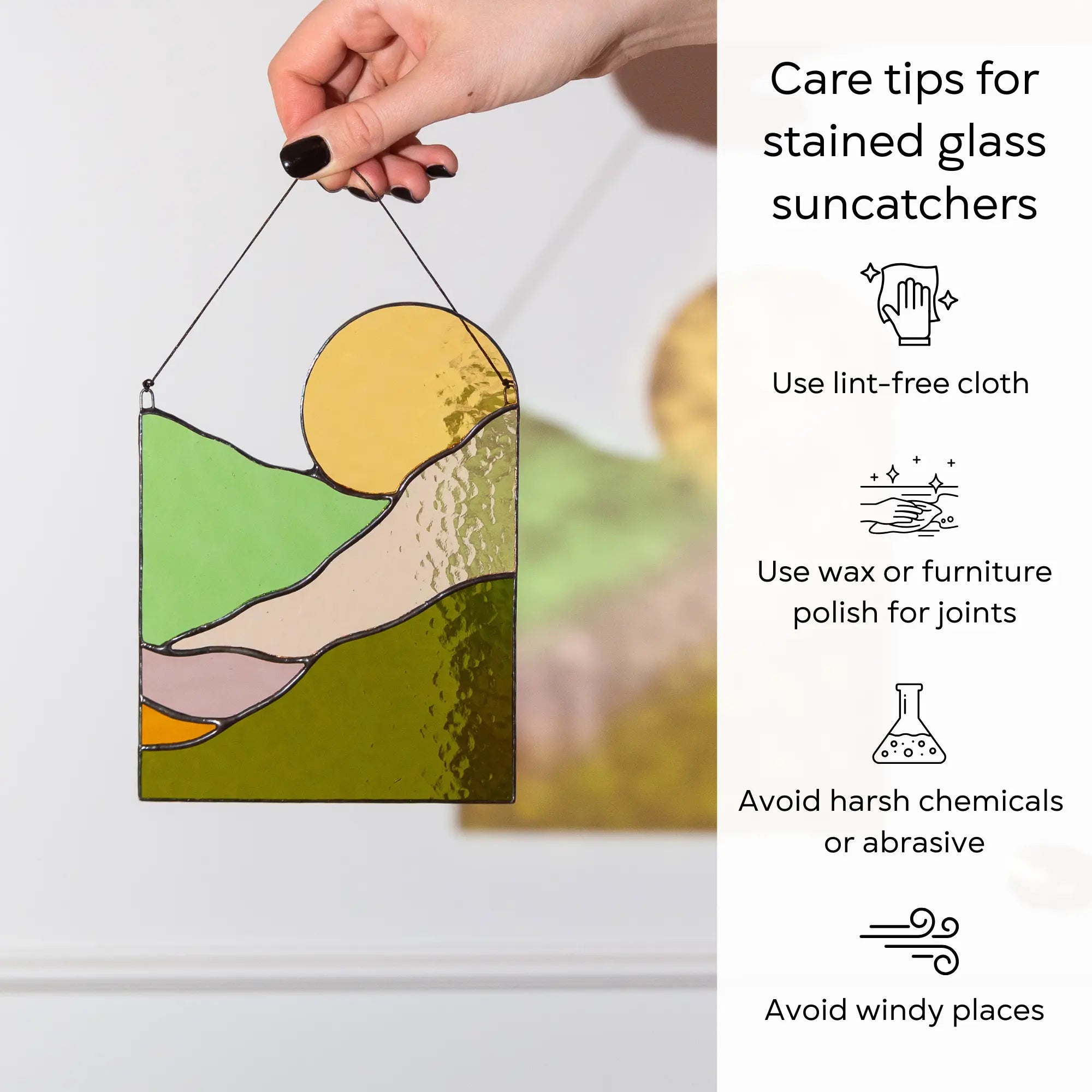 A hand holding a sun landscape stained glass suncatcher next to essential care and cleaning tips: use lint-free cloth, polish joints with car wax or furniture polish, avoid harsh chemicals, and windy places.