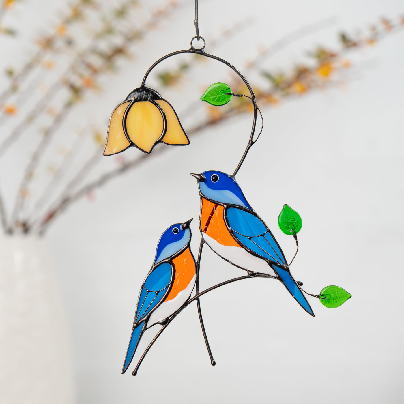 Two stained glass bluebirds sit under a yellow flower with green leaves, hanging as a window decoration
