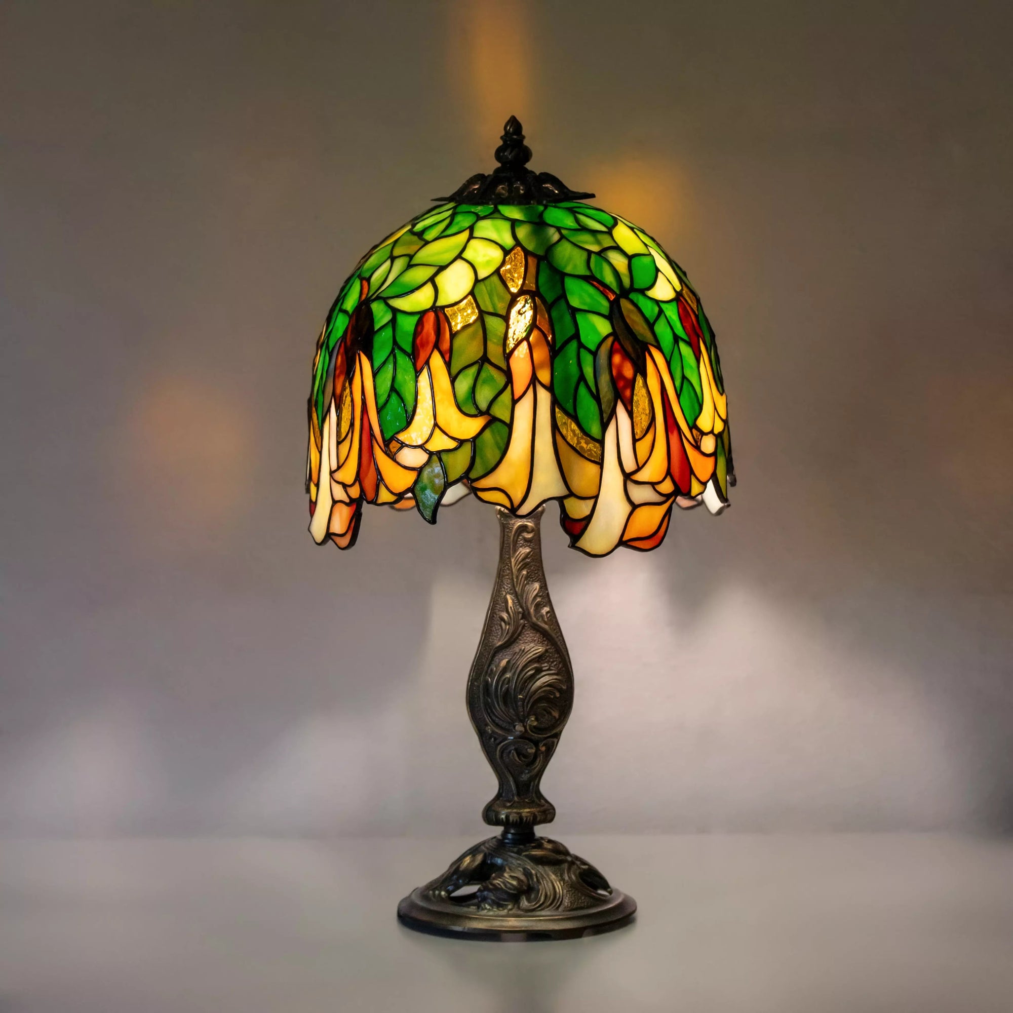 Vintage stained glass Tiffany table lamp with green leaves and yellow flowers glowing warmly on ornate metal base.