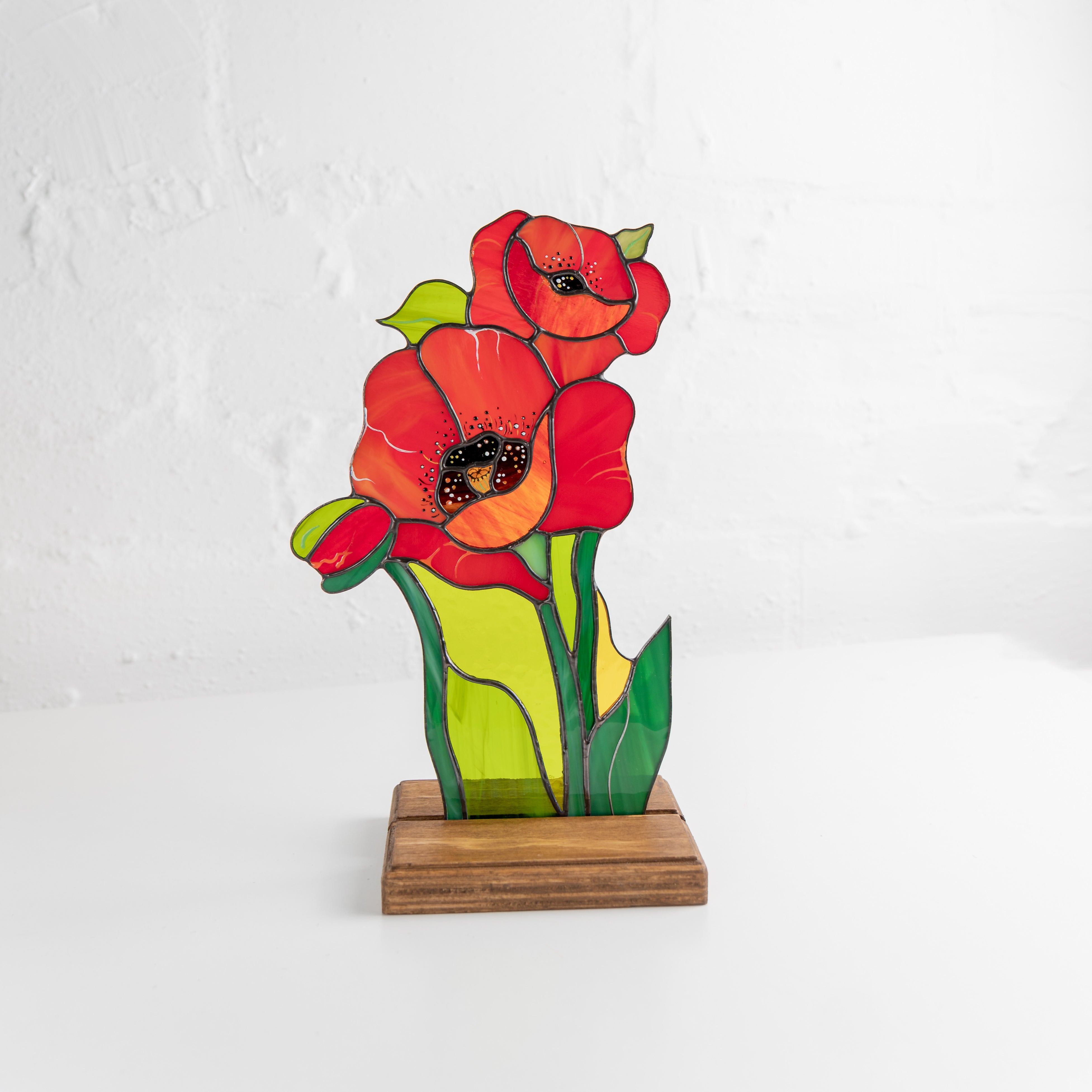 Stained glass poppy flower suncatcher in a wooden stand, set against a white wall background.