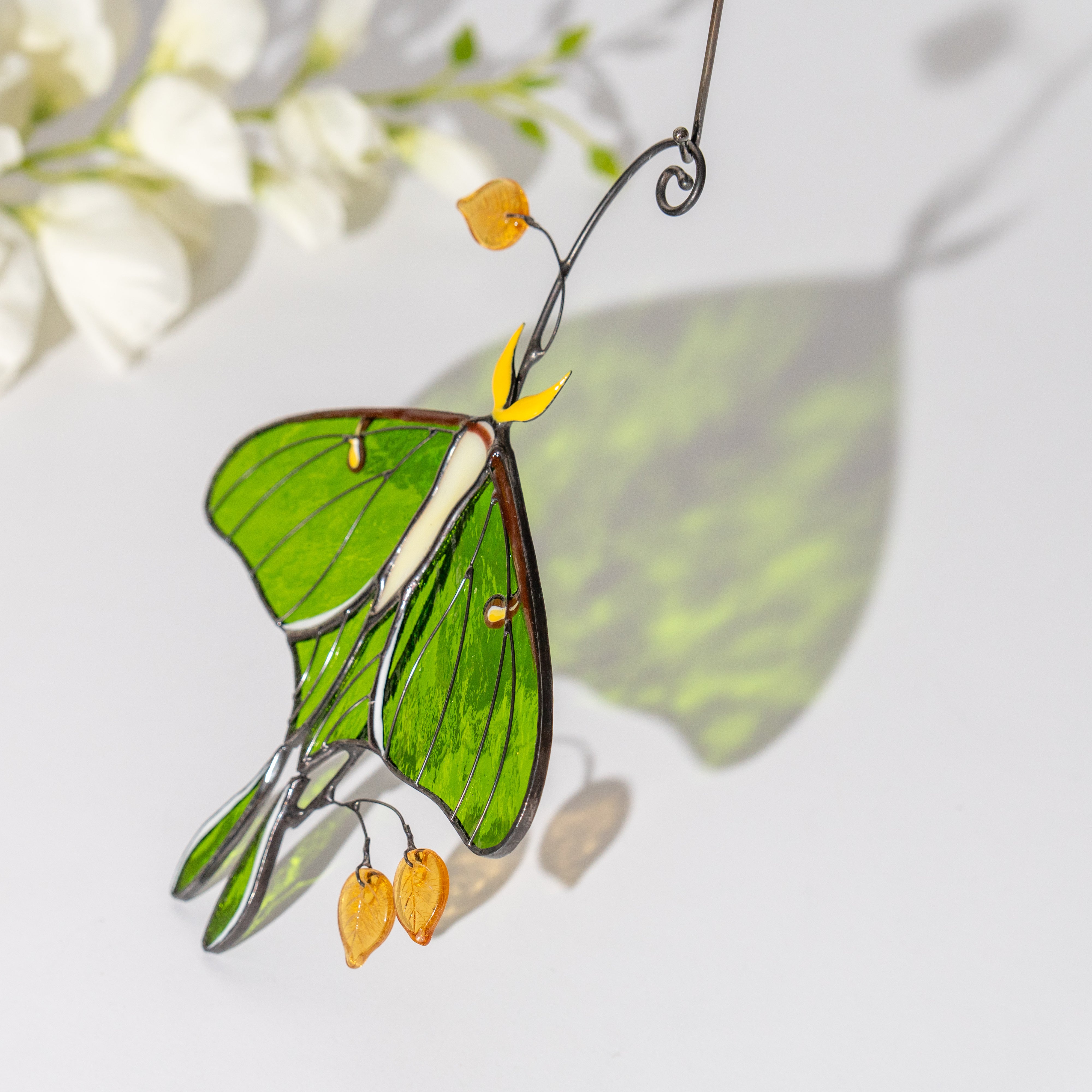 Luna moth suncatcher casting a green shadow on a white wall, with delicate floral accents.