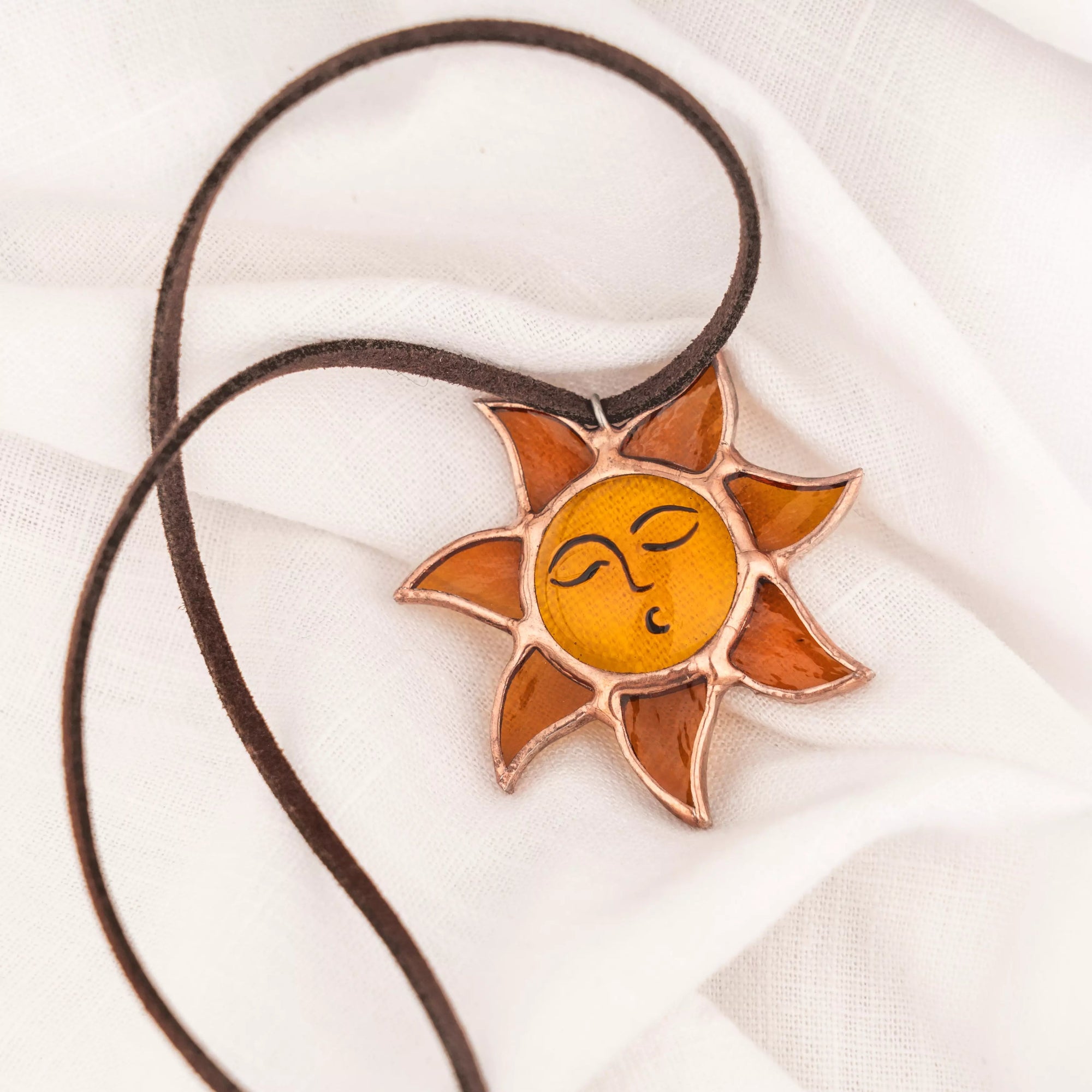 Stained glass Sun pendant with closed eyes on brown cord, lying on textured white cloth with copper foil outline	