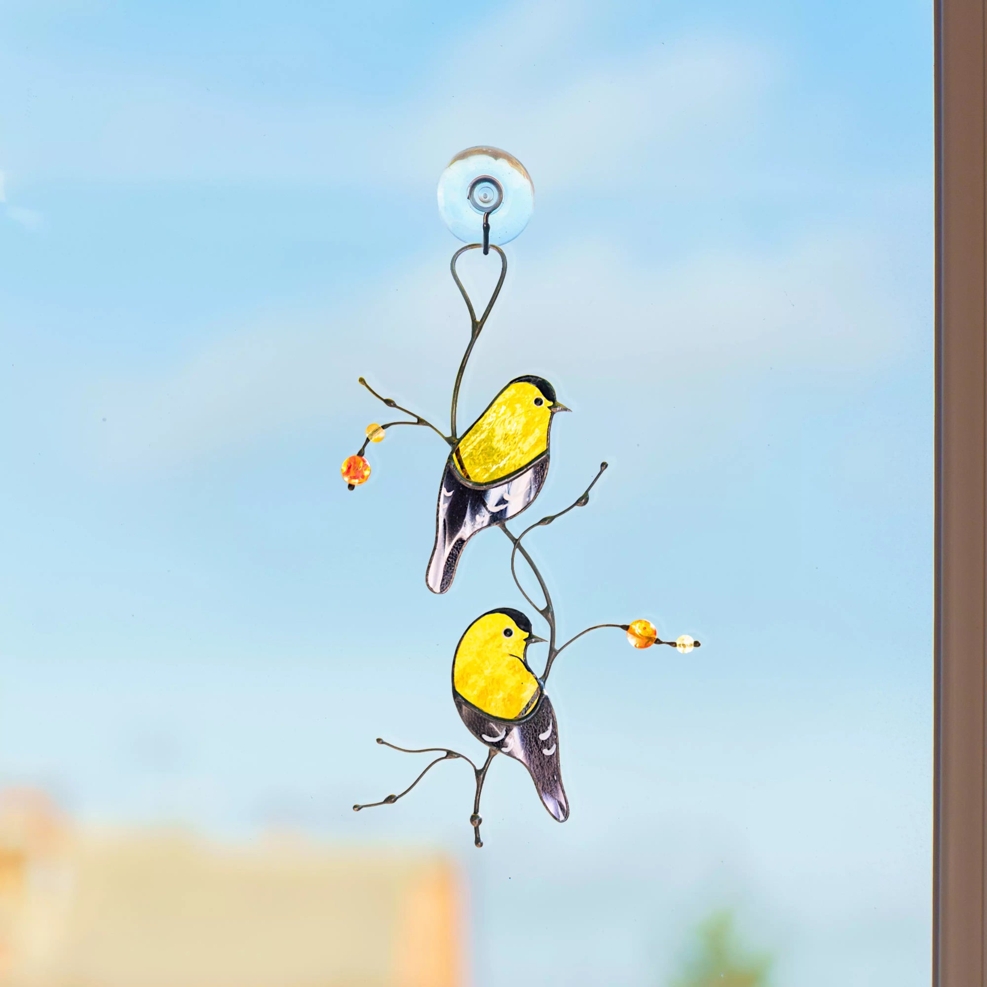 Pair of stained glass goldfinches displayed on a window using a suction cup, with a clear blue sky in the background.