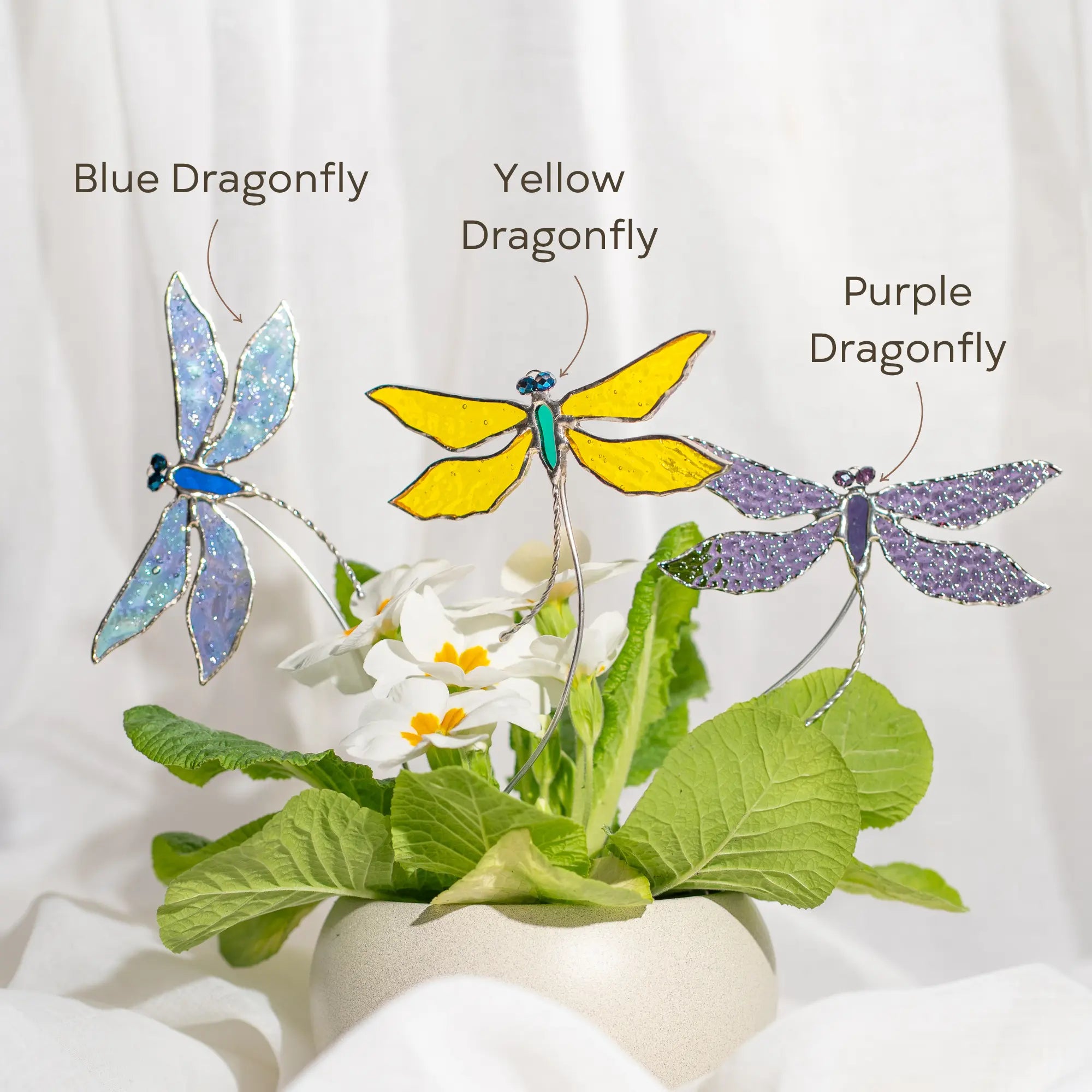 Three dragonfly stained glass plant stakes in blue, yellow, and purple on a white background with green leaves.