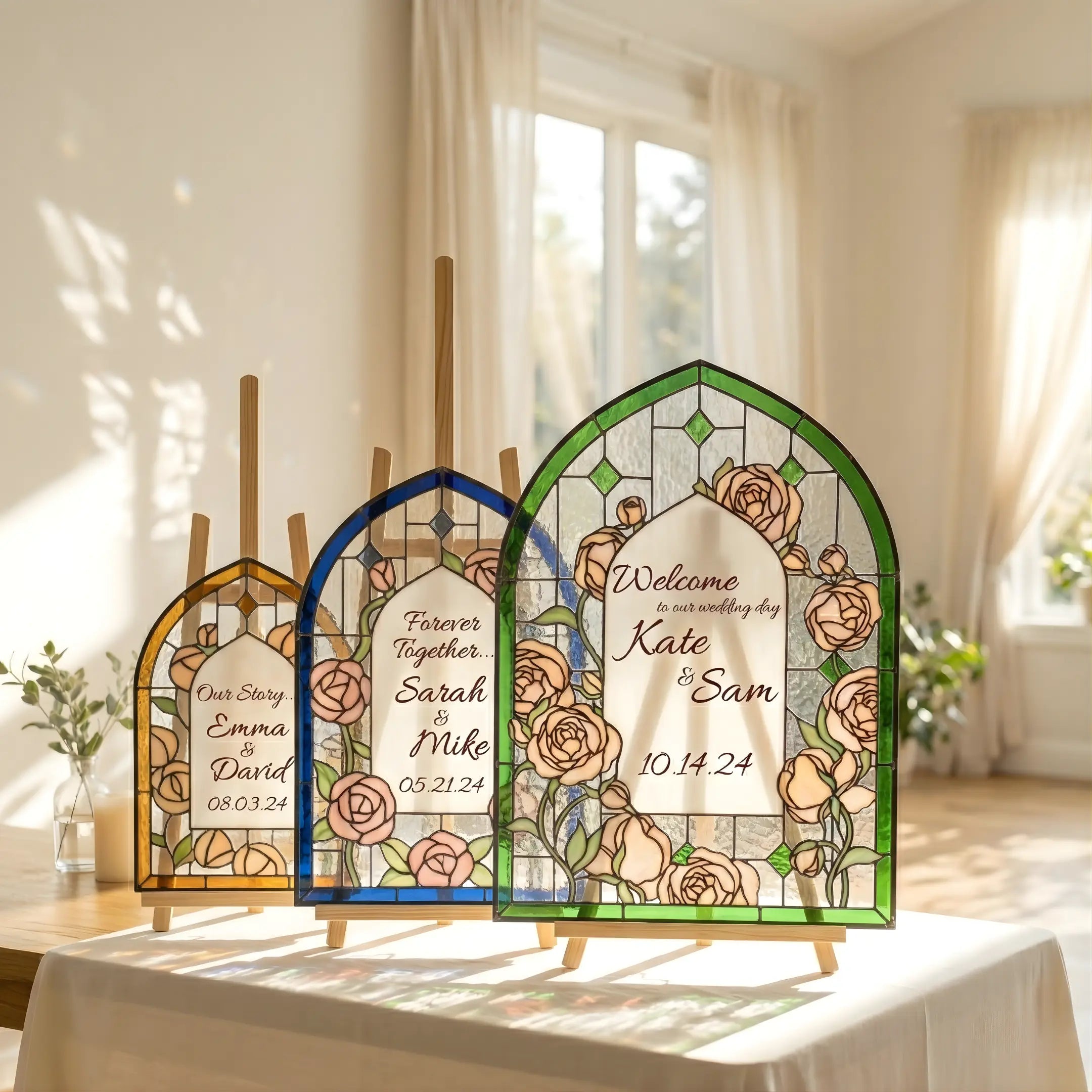 Three arched stained glass wedding sign panels with gold, blue, and green borders displayed on mini easels in a sunlit room.