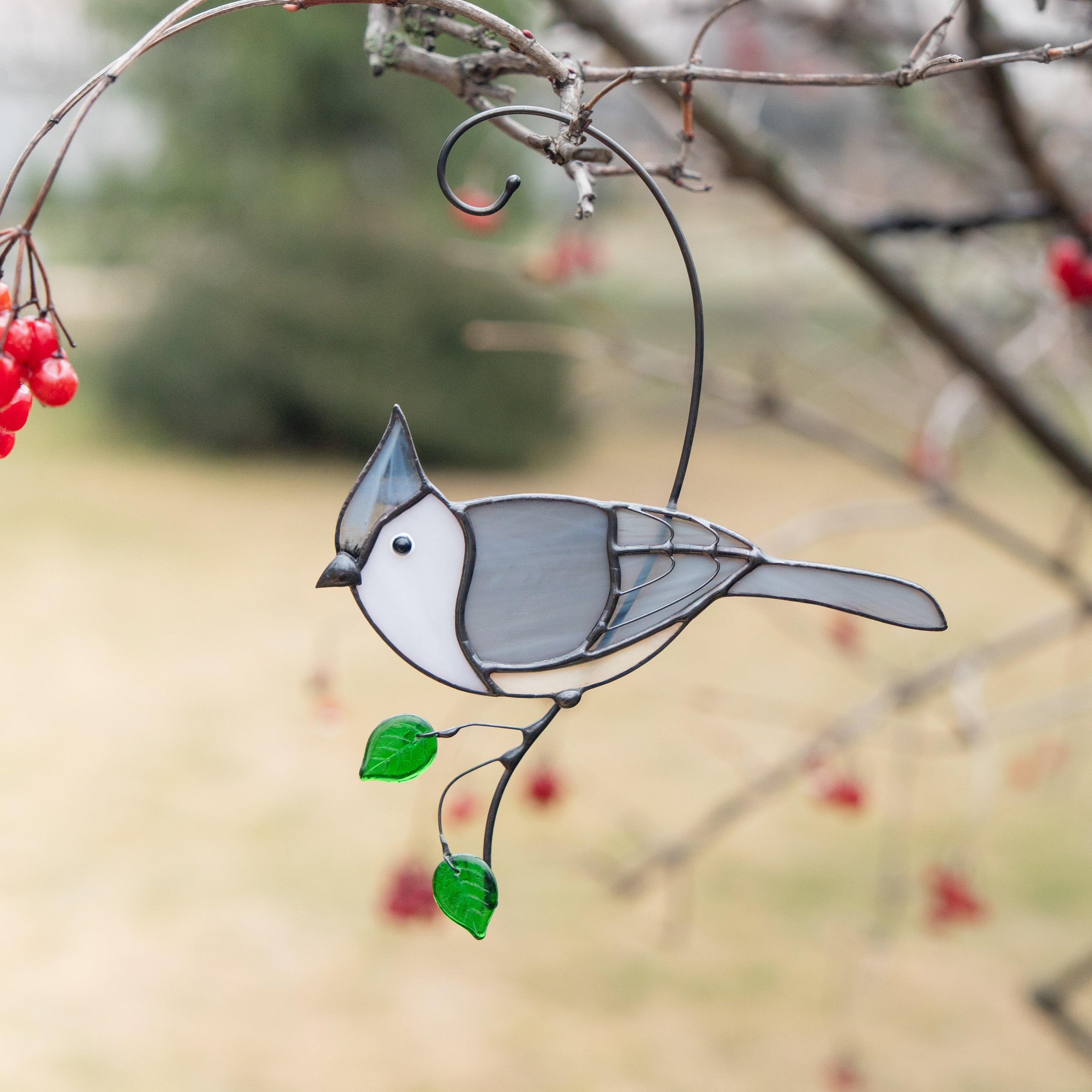 Stained glass tufted titmouse hanging on a tree branch with green leaves and red berries in the background.