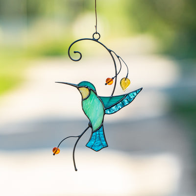 Stained glass hummingbird in turquoise and aqua tones with amber beads, suspended in sunlight against a soft path backdrop