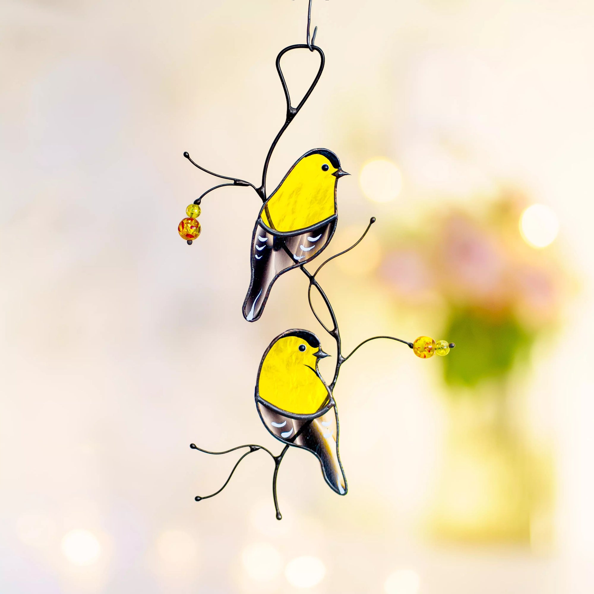 The goldfinch stained glass panel hanging against a soft-focus background of spring flowers, showcasing its vibrant colors.