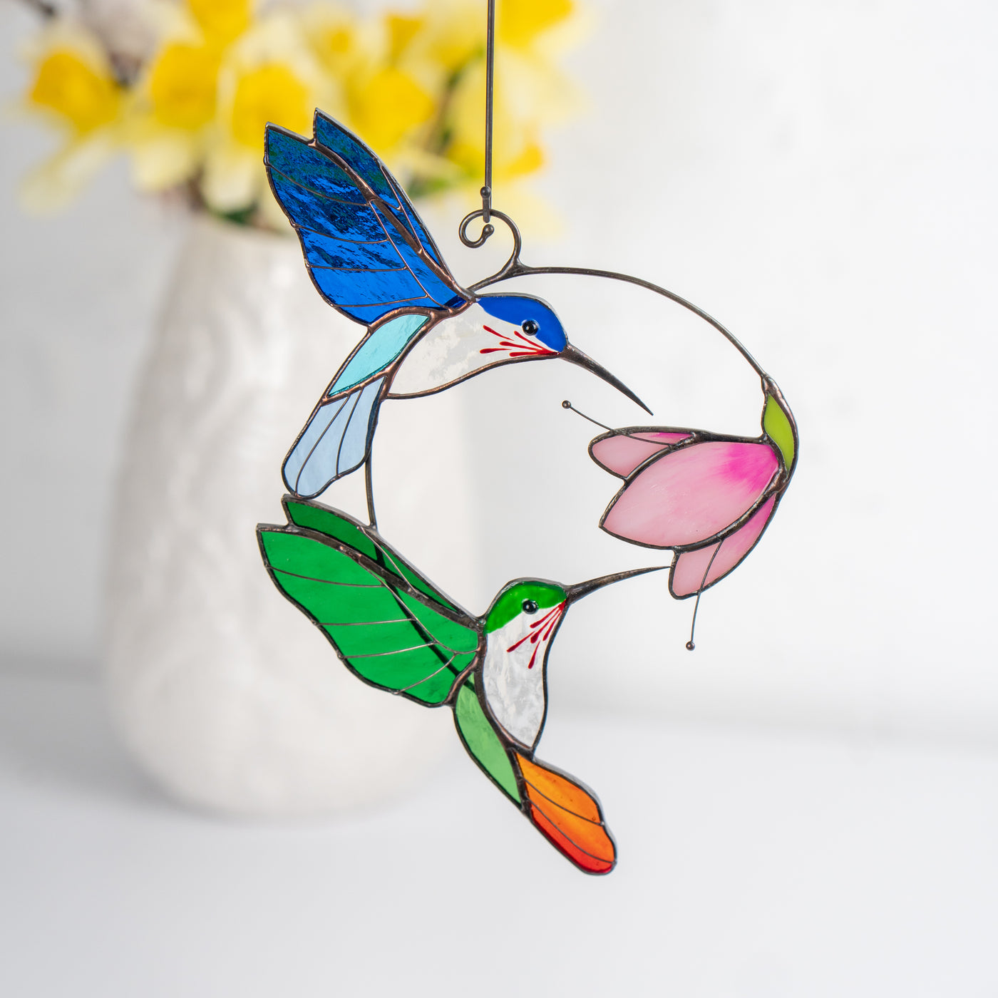 Stained glass suncatcher with two hummingbirds and a pink flower, displayed in front of a vase.