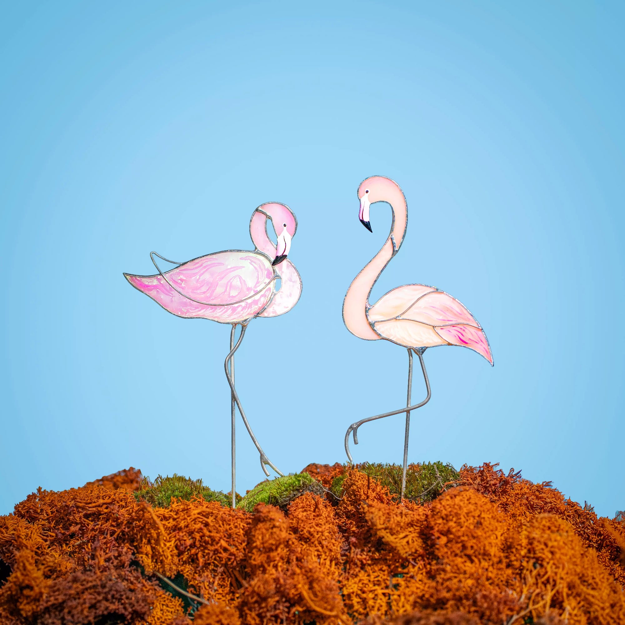 Two pink flamingo stained glass plant stakes on a bed of orange moss with a clear blue sky background.