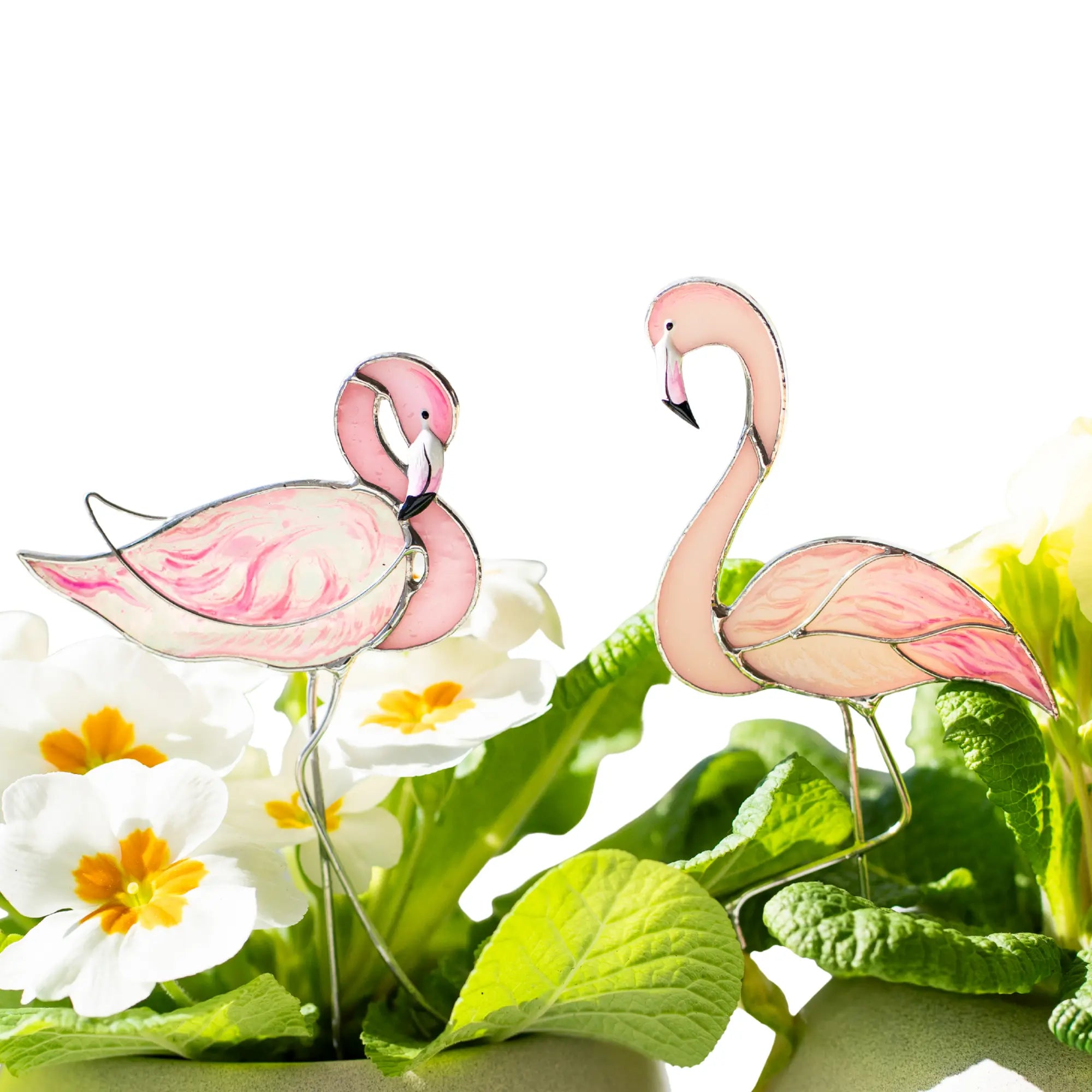Two pink flamingo stained glassplant stakes with greenery and flowers on a white background