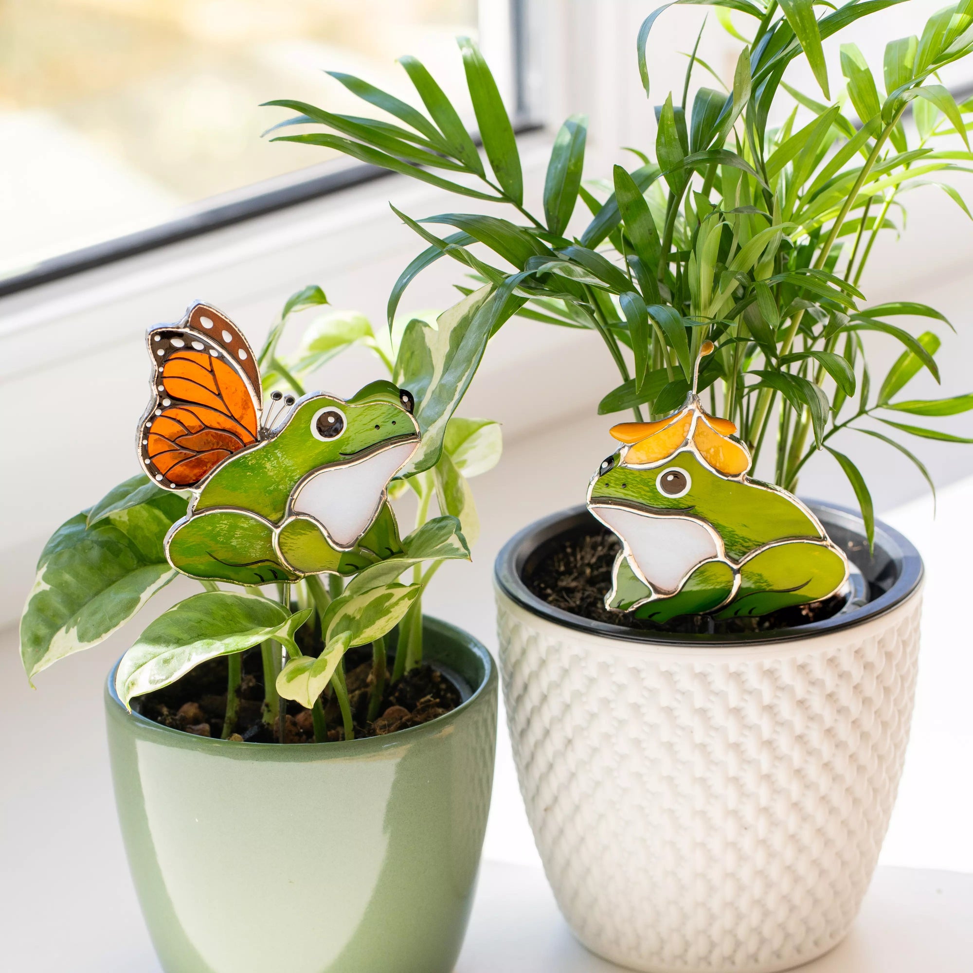 Potted plants with stained glass frog and butterfly plant stakes on a light background.