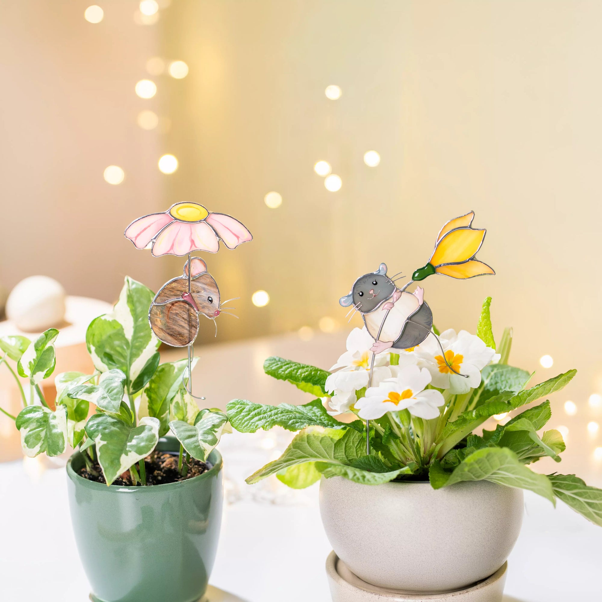 Two potted plants with stained glass mice and flower plant stakes against a warm, blurred background.