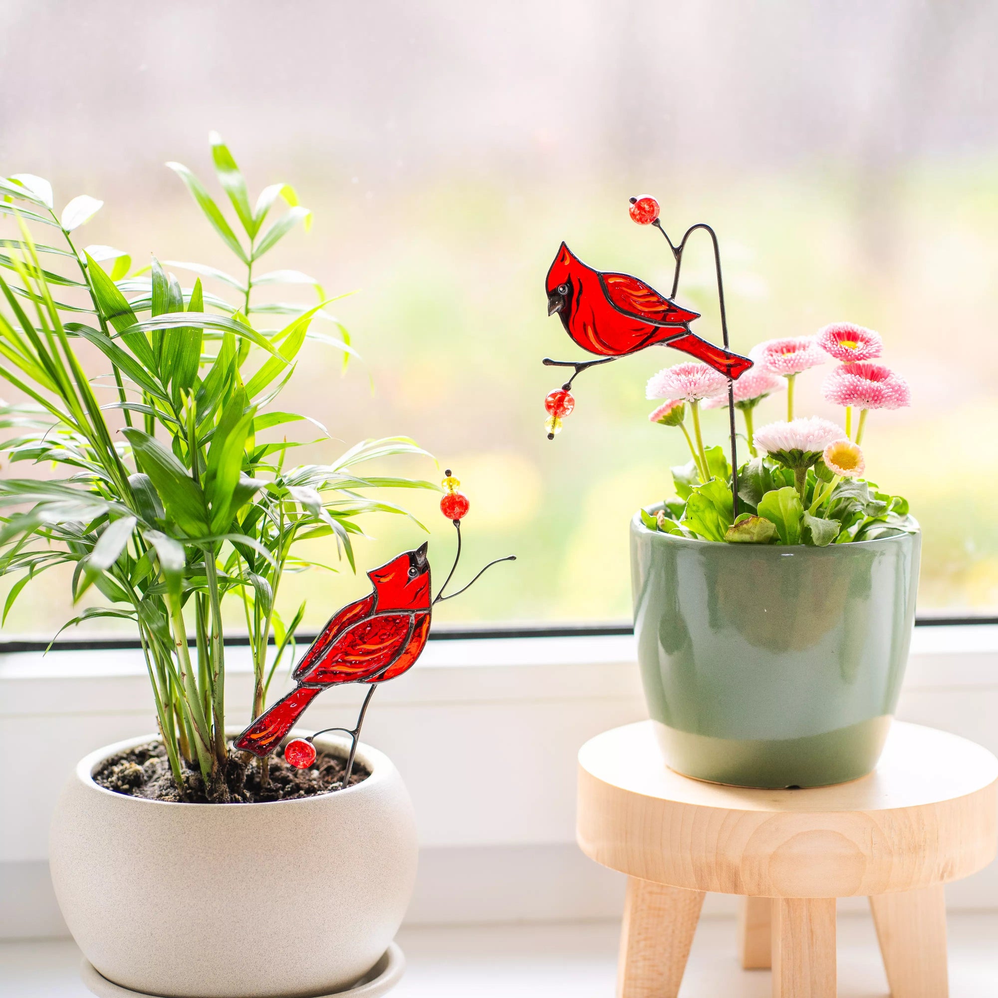 Two potted plants with red cardinal staines glass plant stakes decorations on a windowsill.