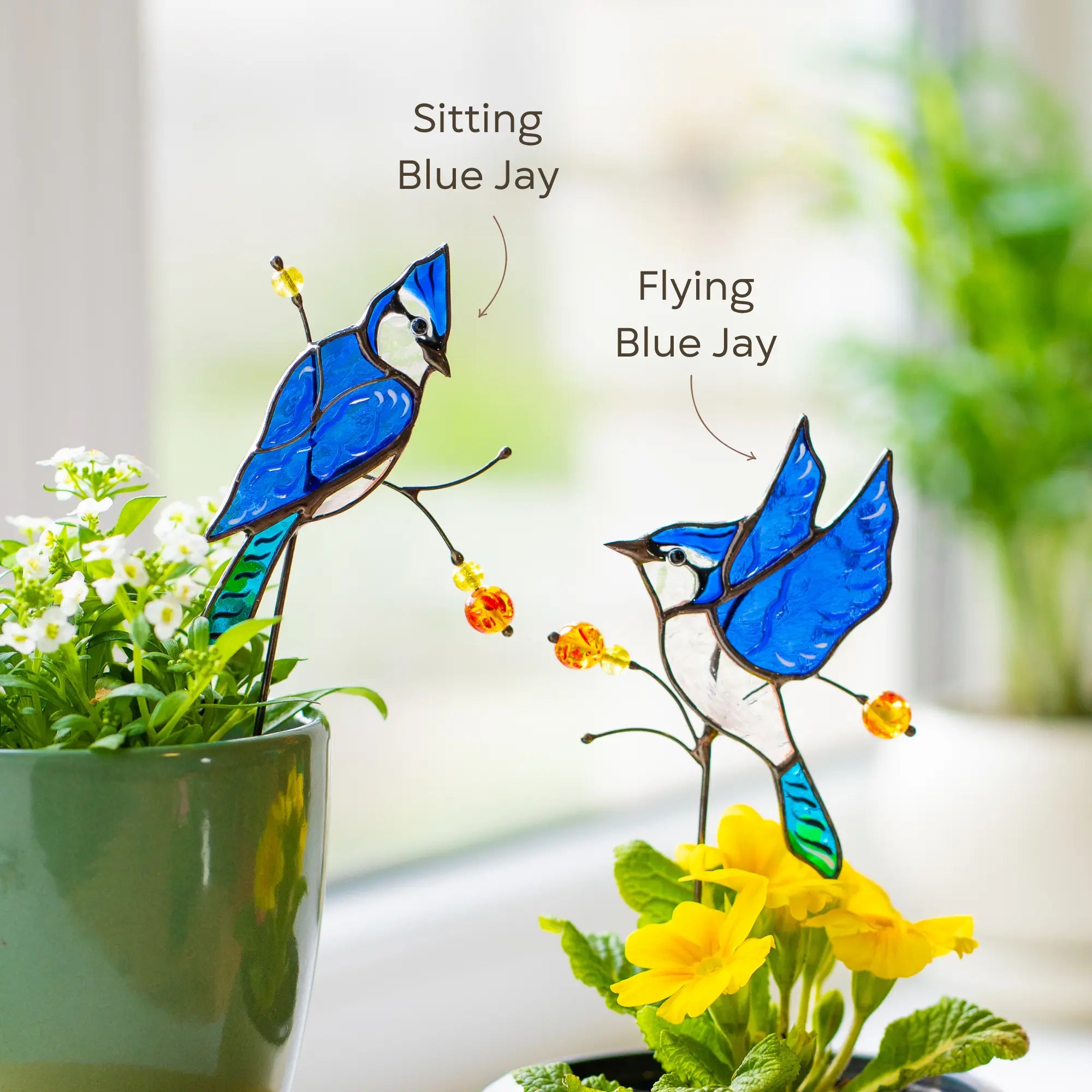 Two stained glass blue jay plant stakes on branches with flowers, one sitting and one flying, in a home setting.
