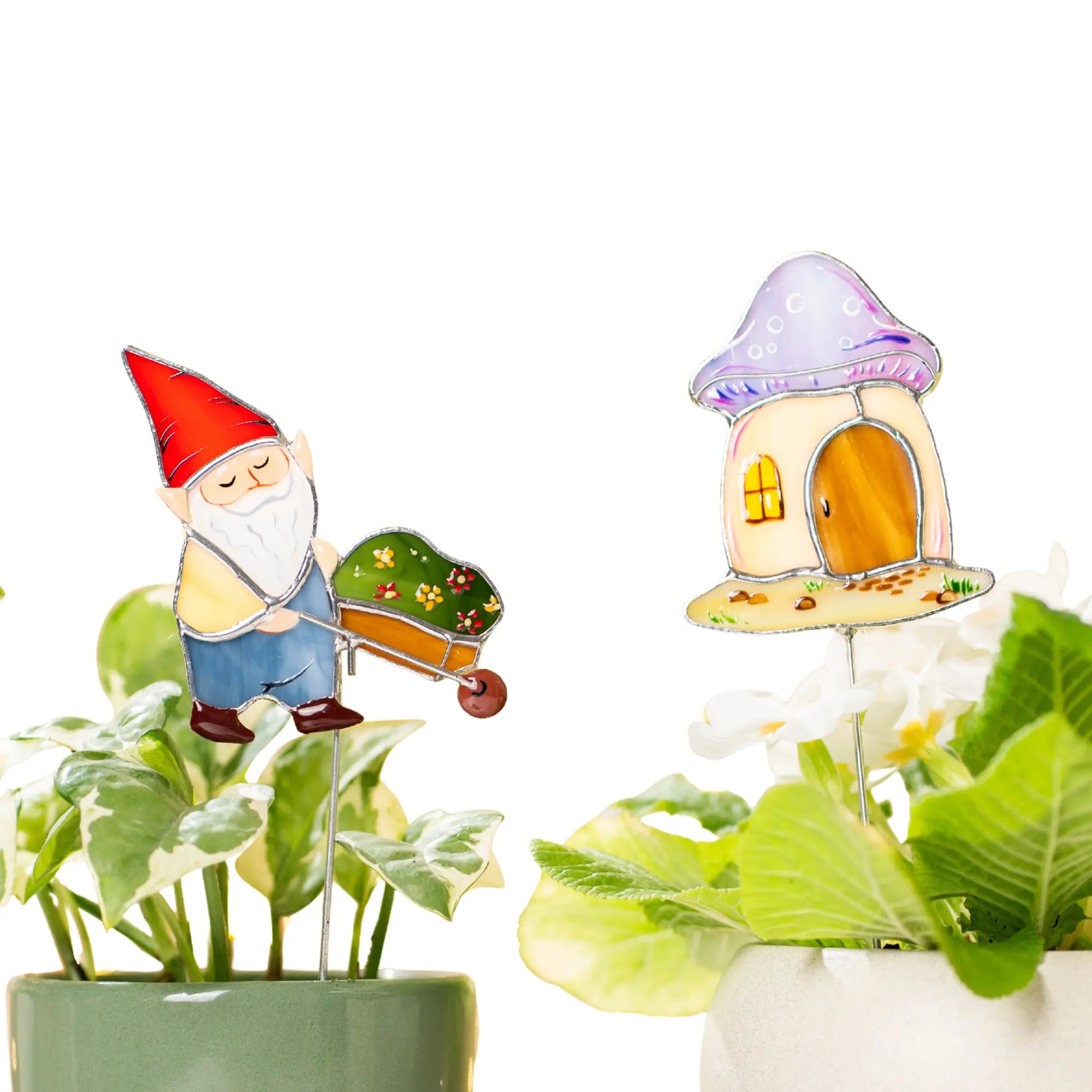 Two stained glass plant stakes, one of a gnome with a red hat pushing a flower-filled wheelbarrow and one of a mushroom-shaped house with a purple cap, are placed in two separate potted plants against a clean white background.