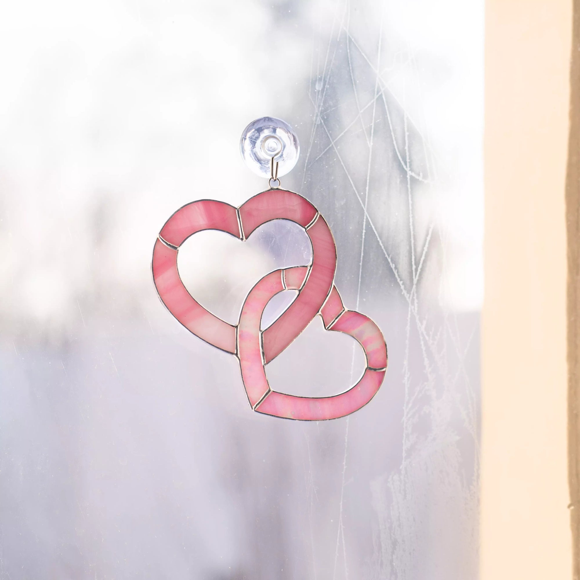 Stained glass pink heart suncatcher attached to window with suction cup, light passing through rose glass.