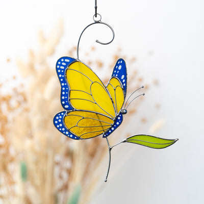 Stained glass butterfly hanging indoors, yellow and blue design with green leaf and soft light background.