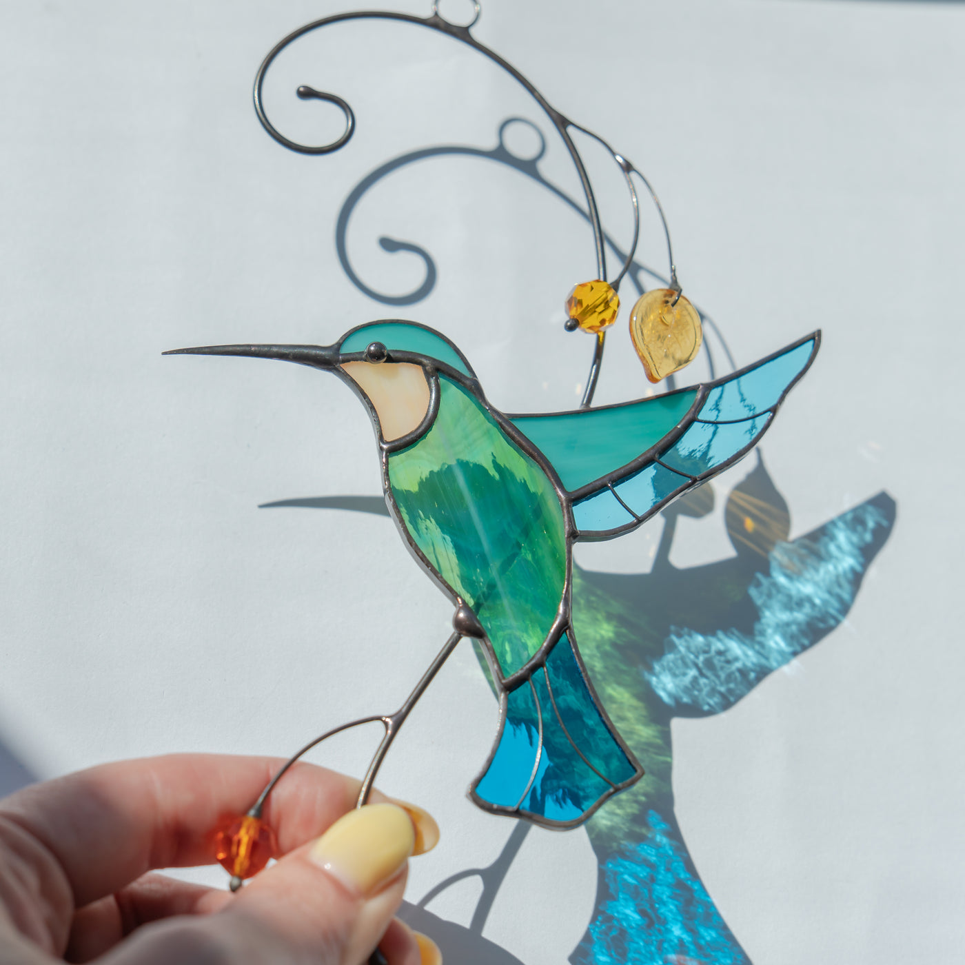 Close-up of a green and blue stained glass hummingbird held in sunlight, casting a colorful shadow on a white background.
