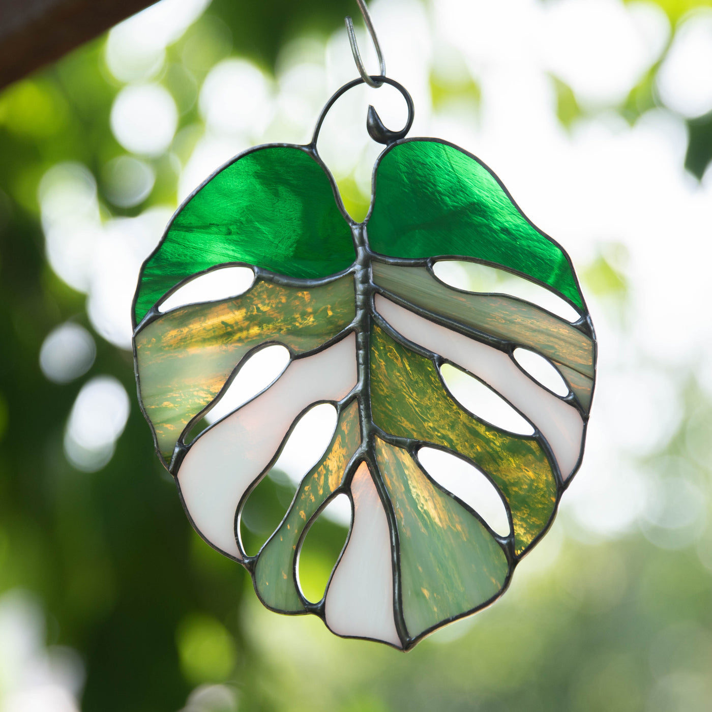 Stained glass monstera suncatcher glowing with sunlight, featuring green, olive, and white glass against leafy background.

