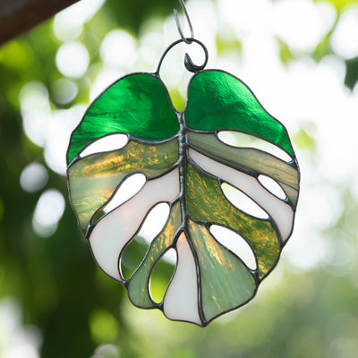 Stained glass monstera suncatcher glowing with sunlight, featuring green, olive, and white glass against leafy background.

