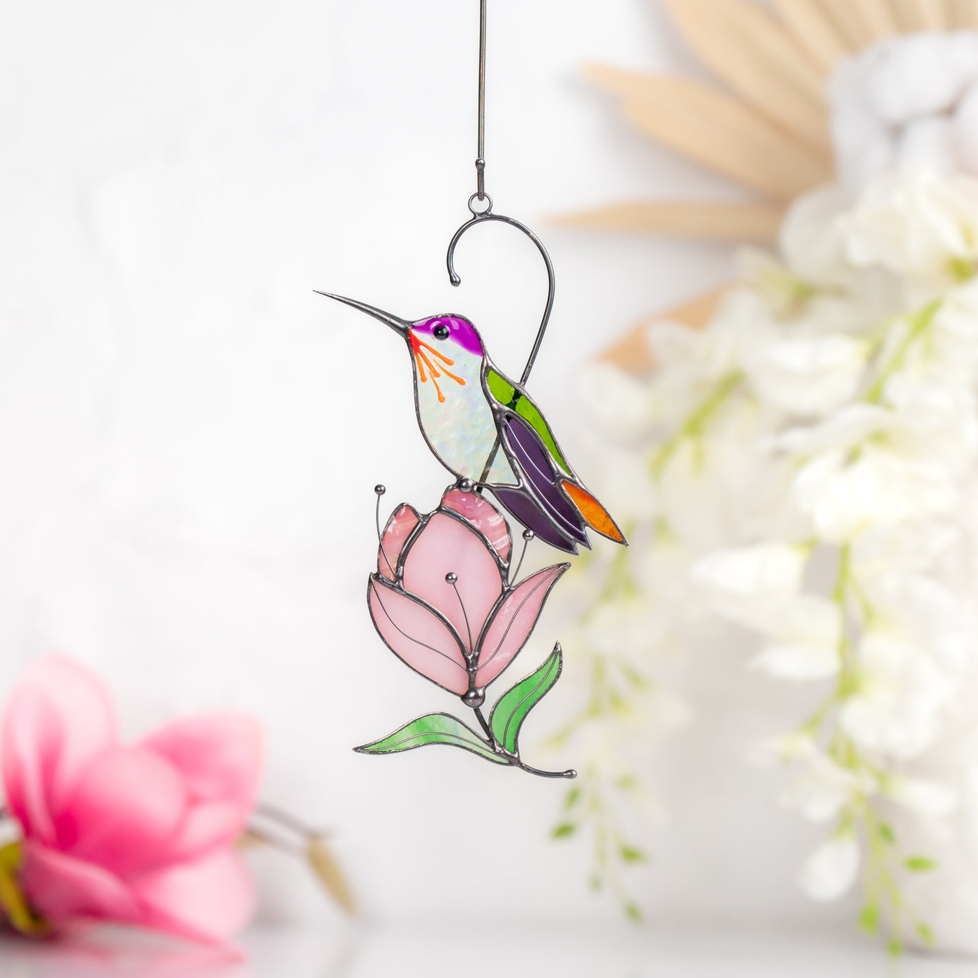 Stained glass hummingbird with purple head and green wings, perched on a soft pink magnolia flower.